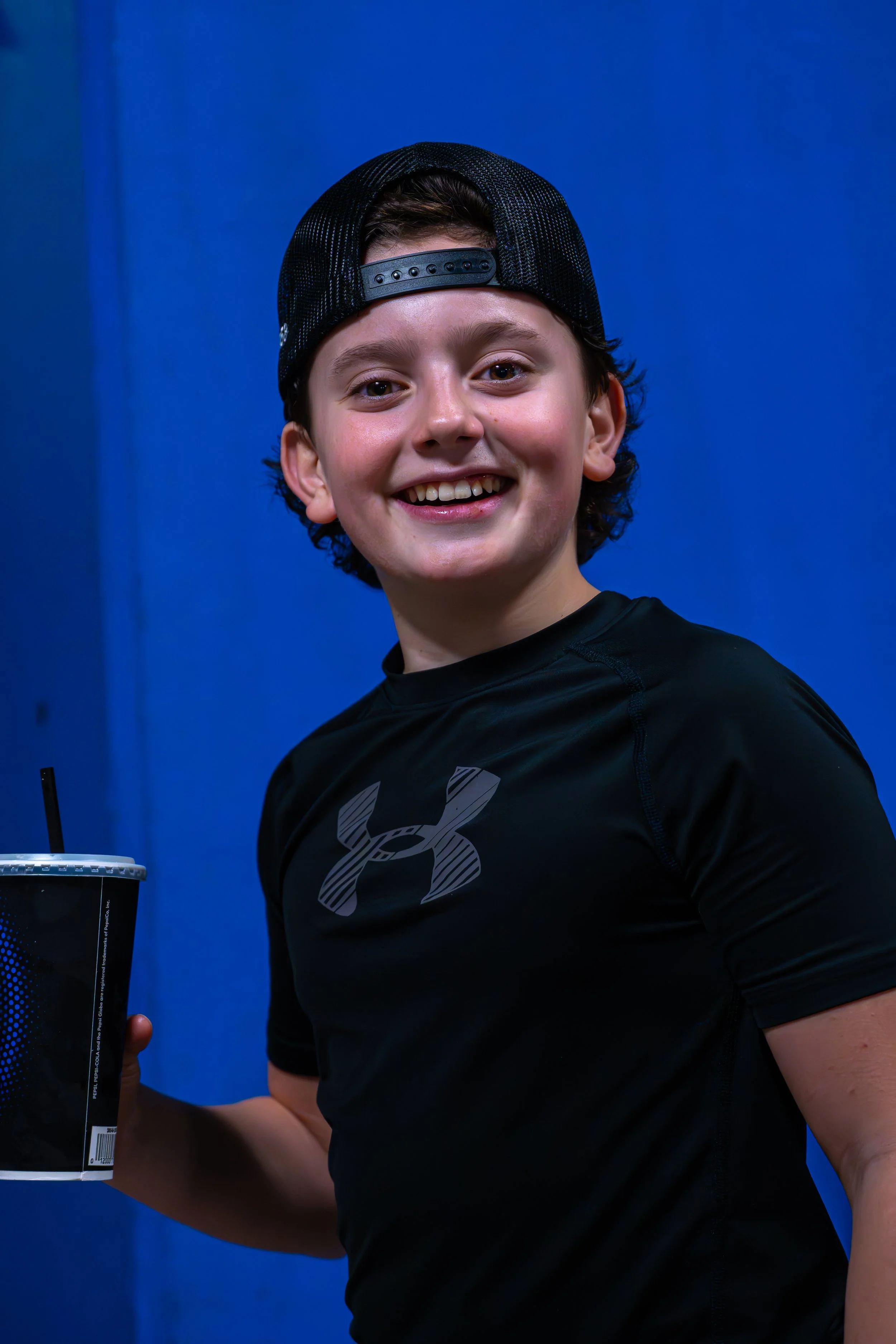 A young boy wearing a black Under Armour shirt and a black baseball cap backwards, smiling and holding a drink cup with a straw against a solid blue background.