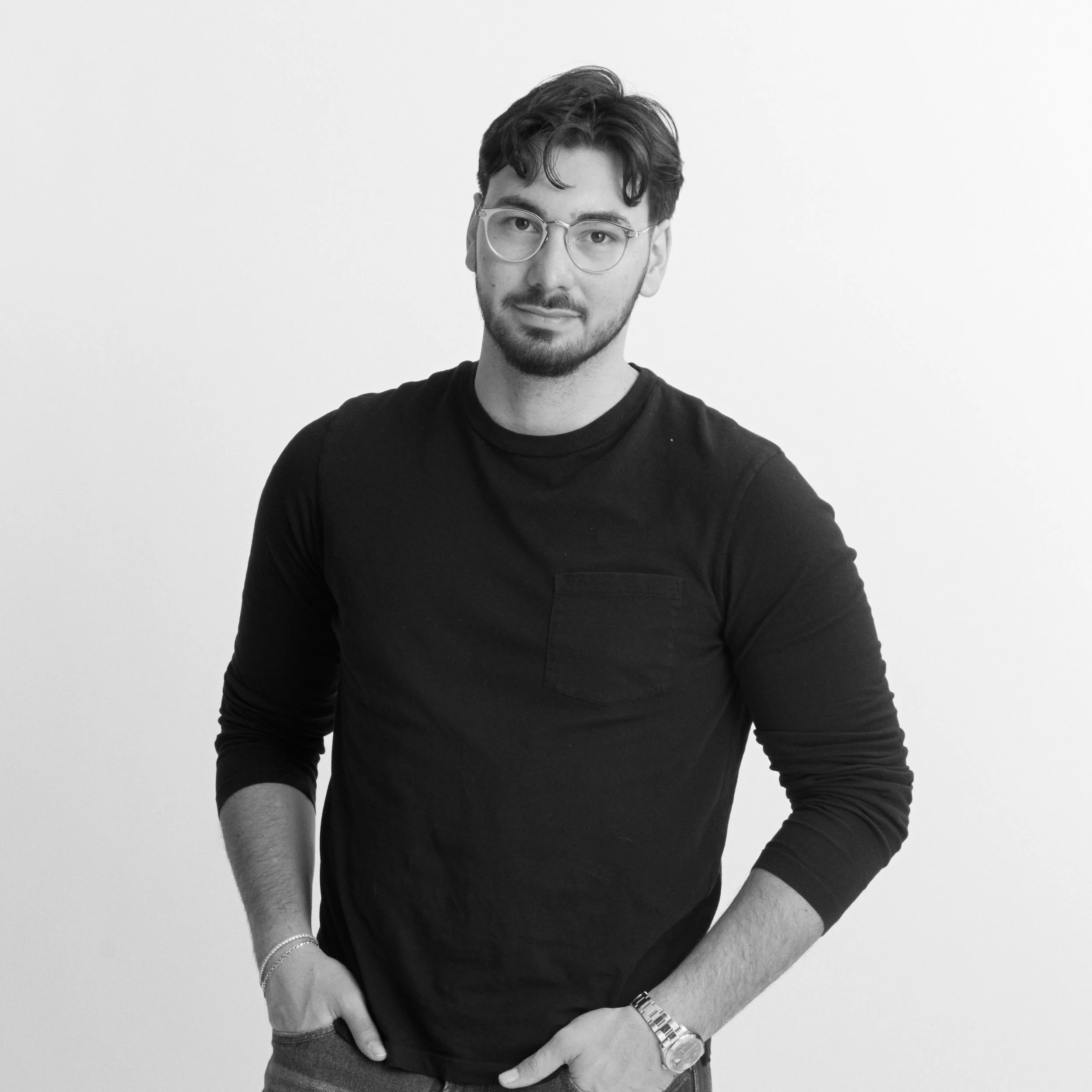 Black and white portrait of a young man with glasses, a beard, wearing a dark long-sleeve shirt, standing against a plain light background.