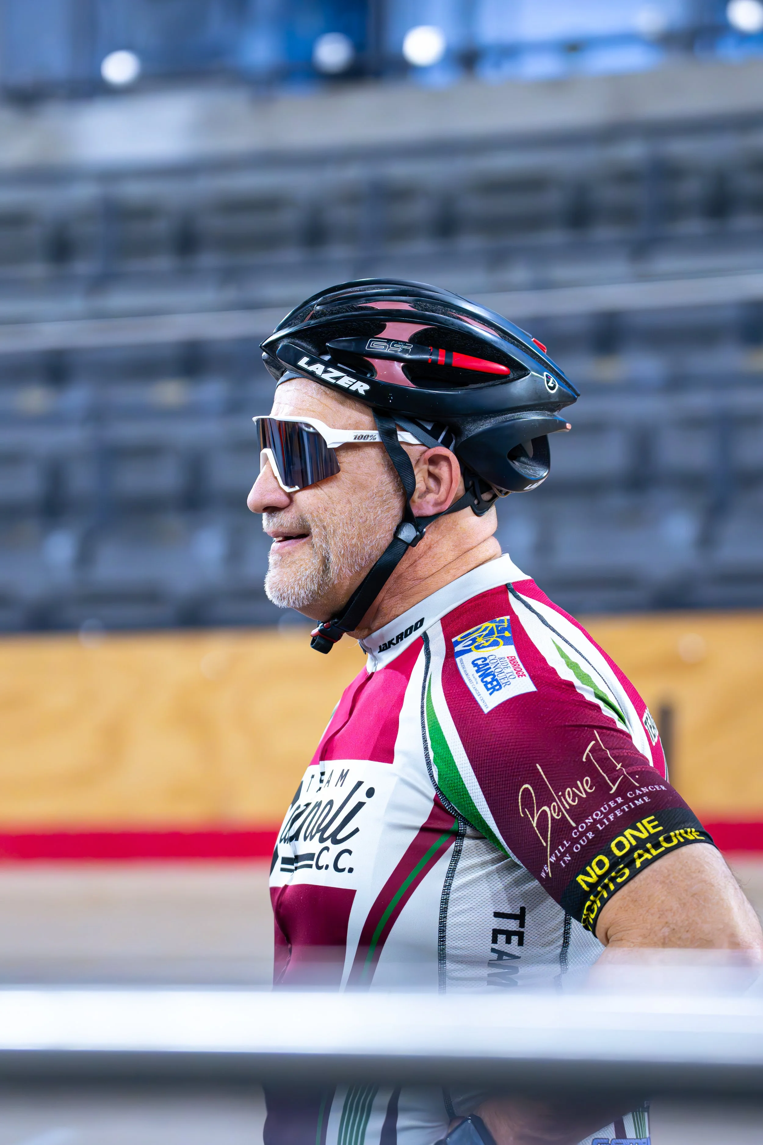 A cyclist wearing a black helmet and white sports sunglasses, dressed in a red, white, and green cycling jersey with sponsor logos and messages, standing in a sports arena with empty bleachers in the background.