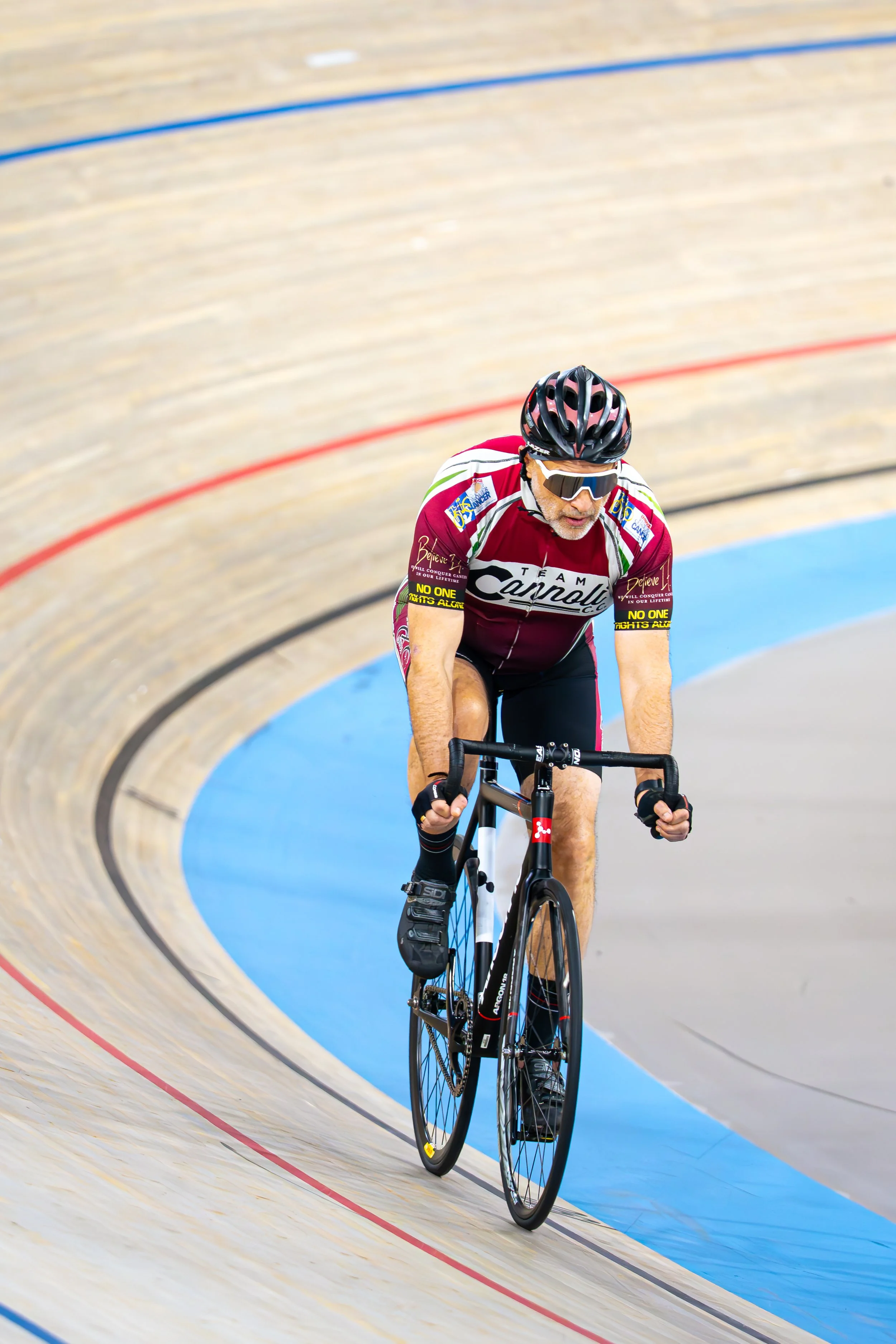 A male cyclist wearing a maroon and white Team Cannondale jersey, sunglasses, and a helmet riding on an indoor velodrome track.