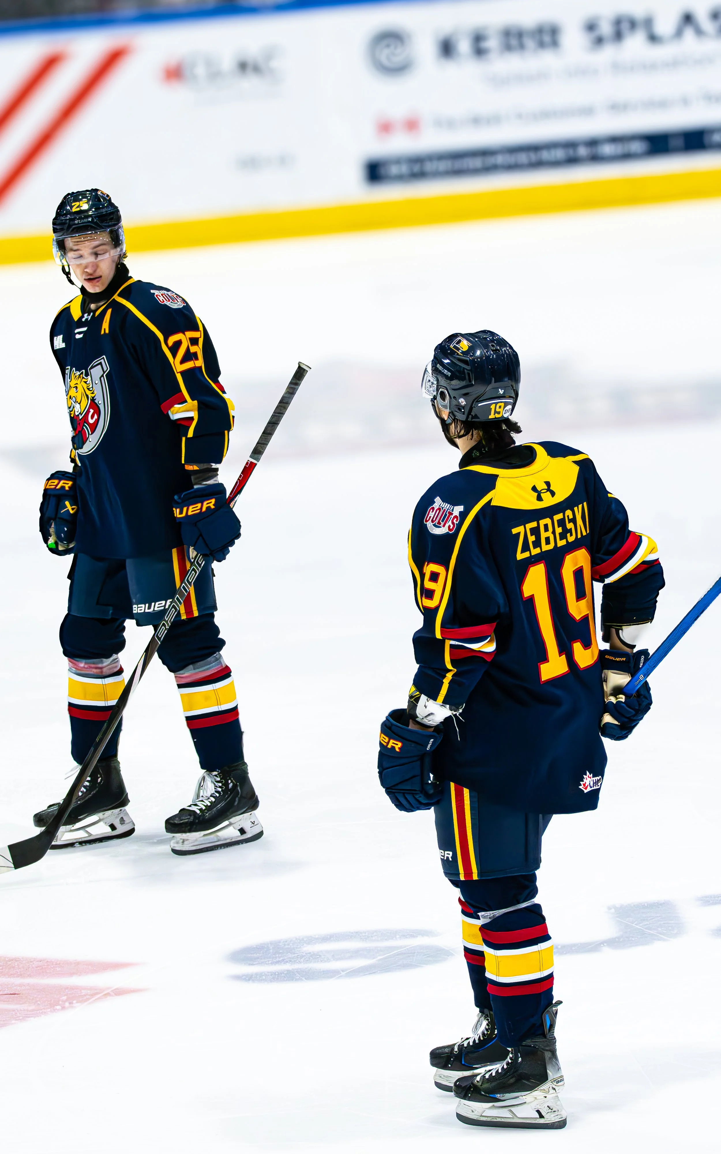 Two ice hockey players in dark blue jerseys with yellow accents on an ice rink. One is standing and looking down while the other is facing away with the name 'Zebeski' and the number 19 on the back.