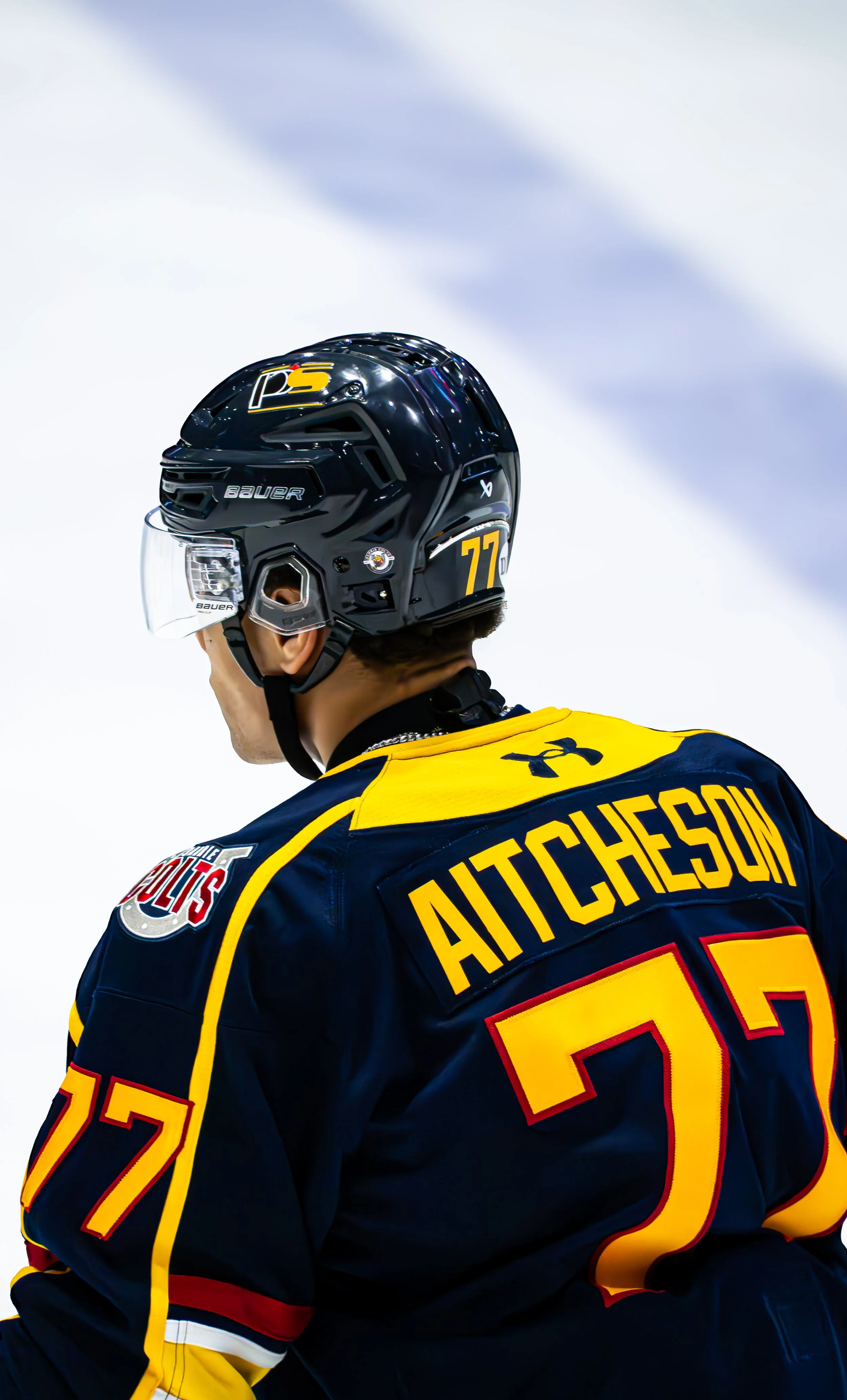 Hockey player wearing a black helmet and a dark blue and yellow jersey with the name 'AITCHESON' and number 77. The player is on ice, with subtle lighting and a blurred background.