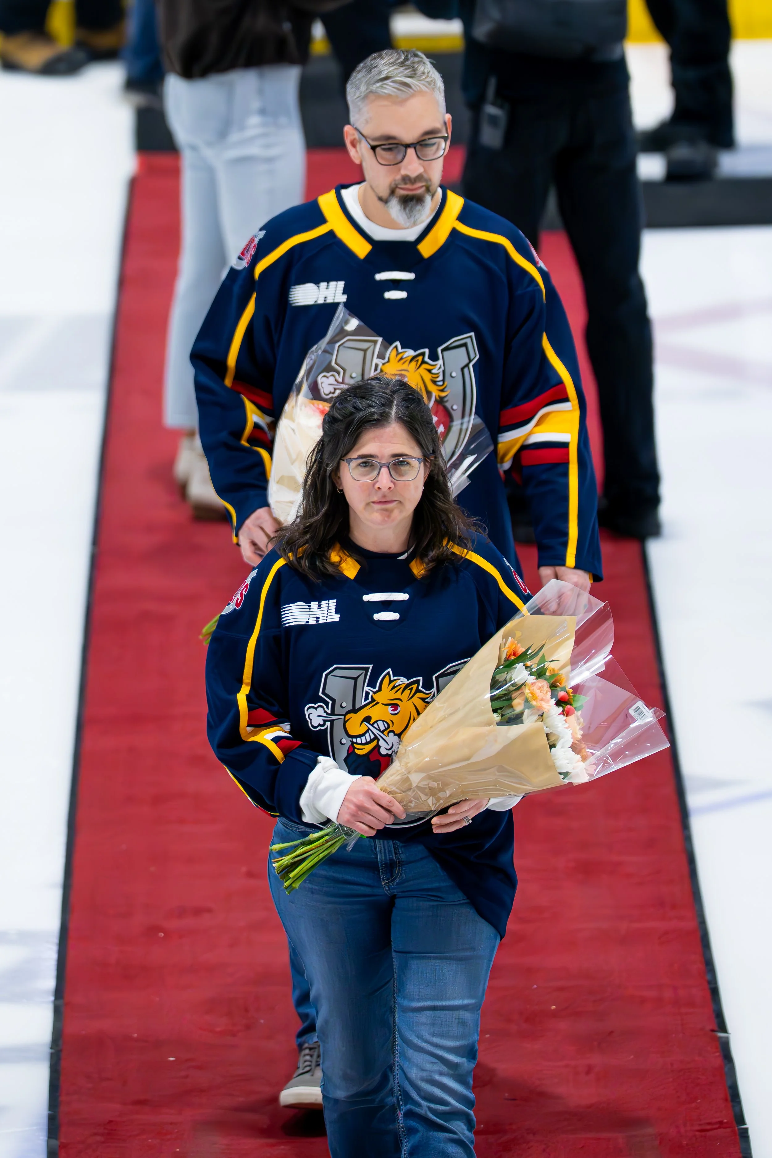 Two people, a man and a woman, wearing hockey jerseys holding bouquets of flowers, walking on a red carpet on an ice rink.