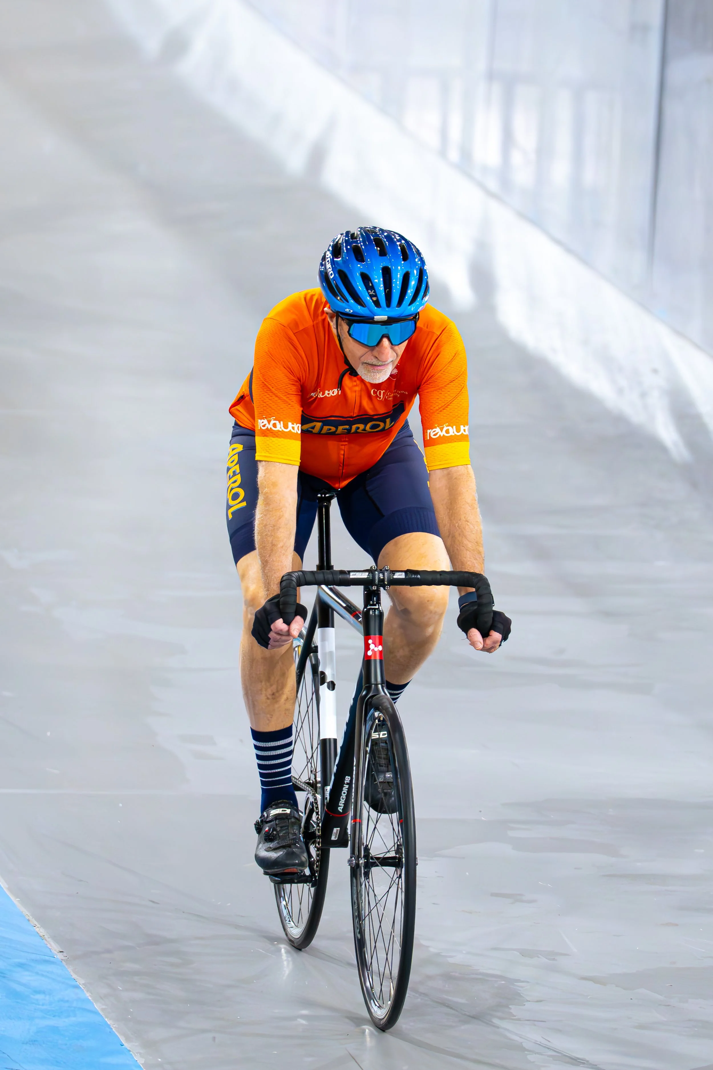 An older man in an orange cycling jersey and dark shorts riding a bicycle indoors on a velodrome track, wearing a blue helmet and sunglasses.