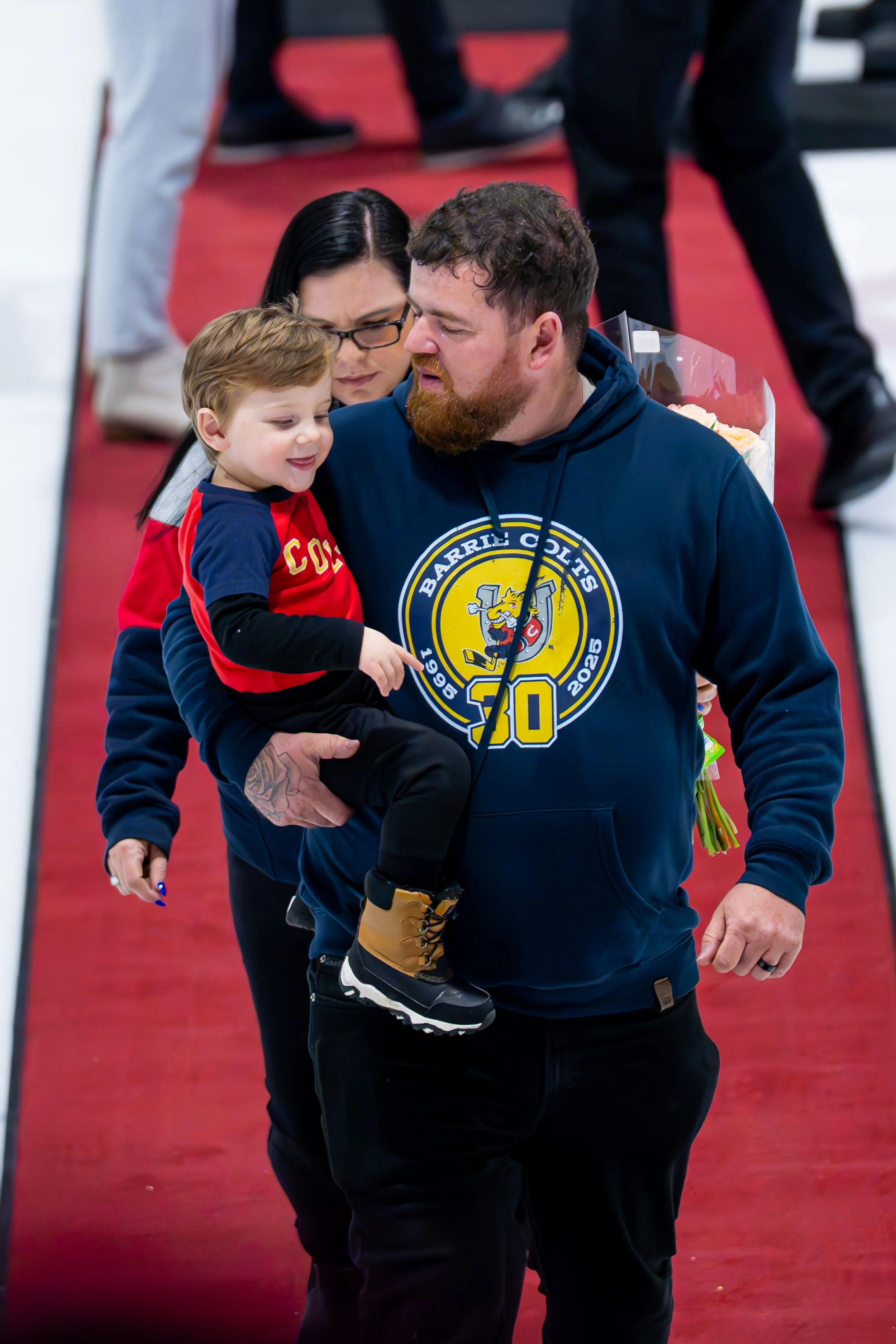 A man with a beard holding a young boy while a woman with black hair looks on at an indoor event, walking on a red carpet.
