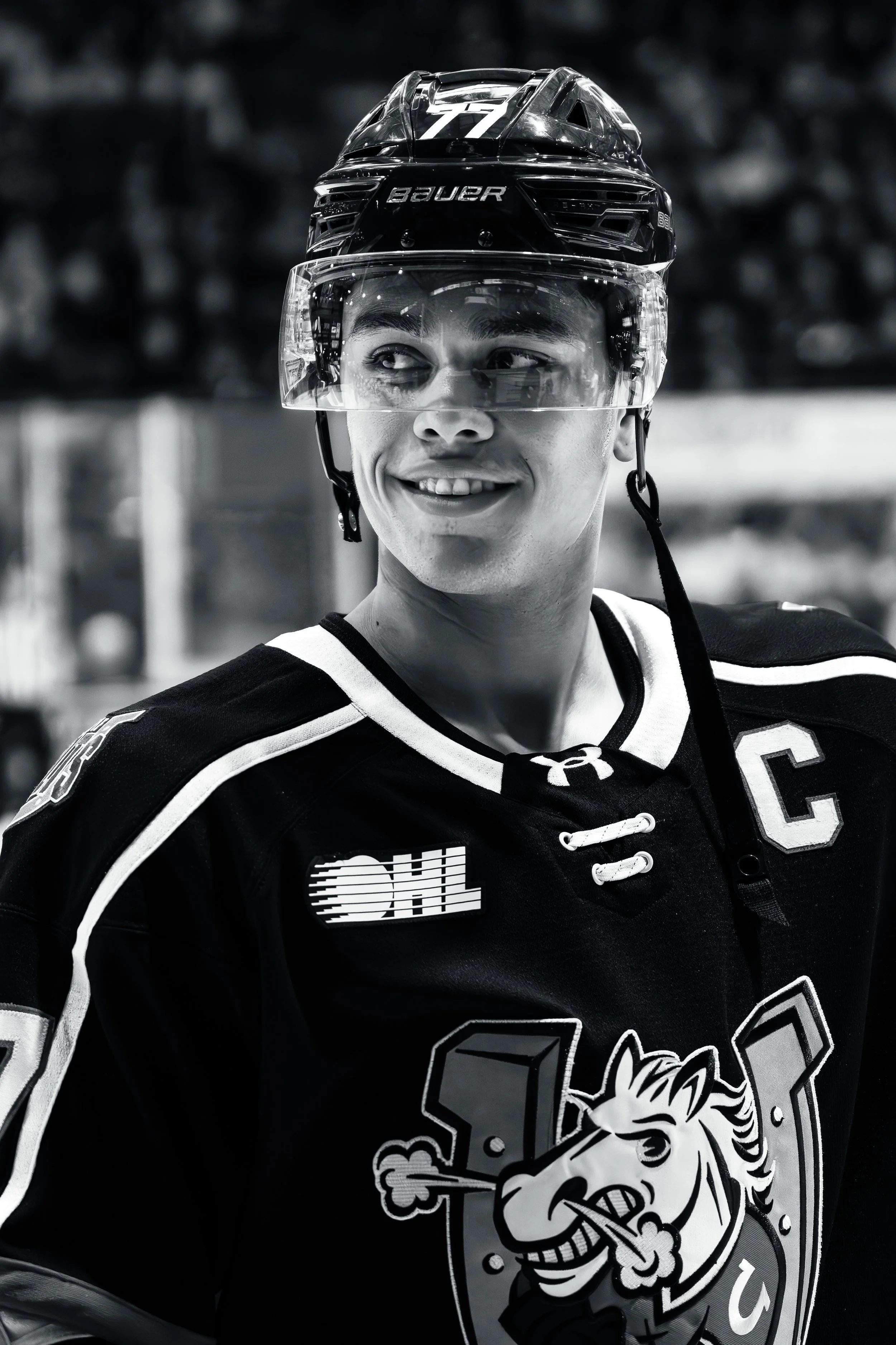 A male ice hockey player wearing a helmet with a visor, a jersey with a unicorn logo, and a Captain's 'C' on his chest, smiling and looking to the side.