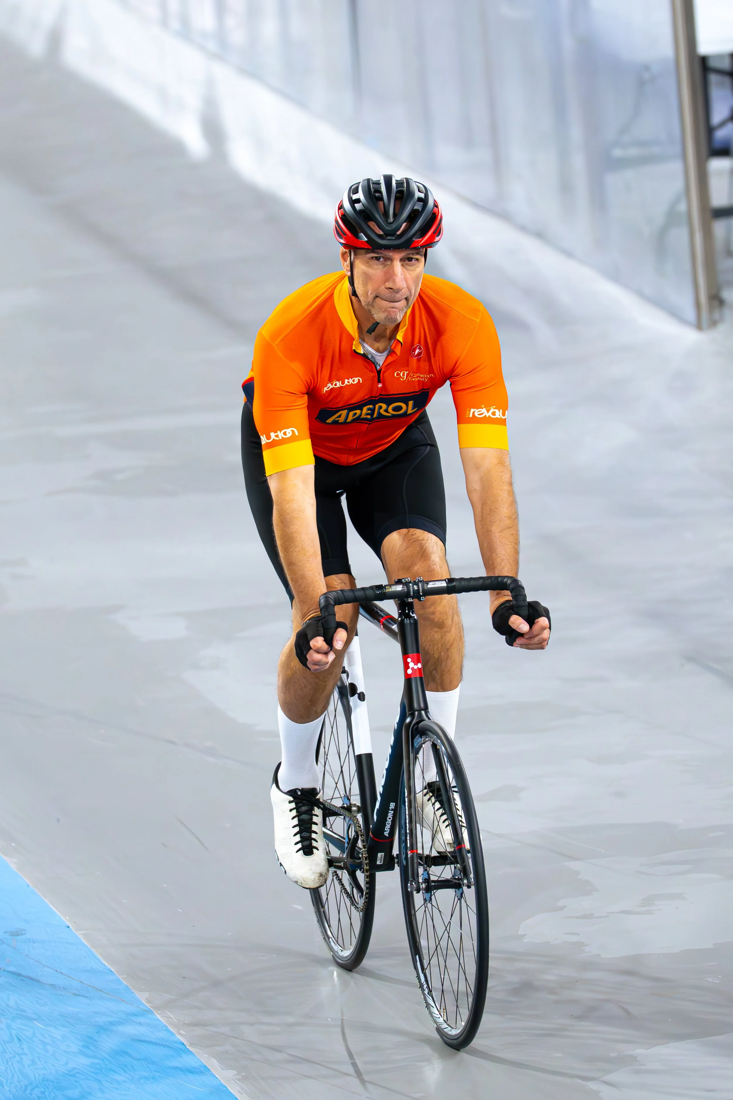 A man wearing a black and red helmet, orange cycling jersey, black cycling shorts, and white socks riding a black track bike indoors on a velodrome.