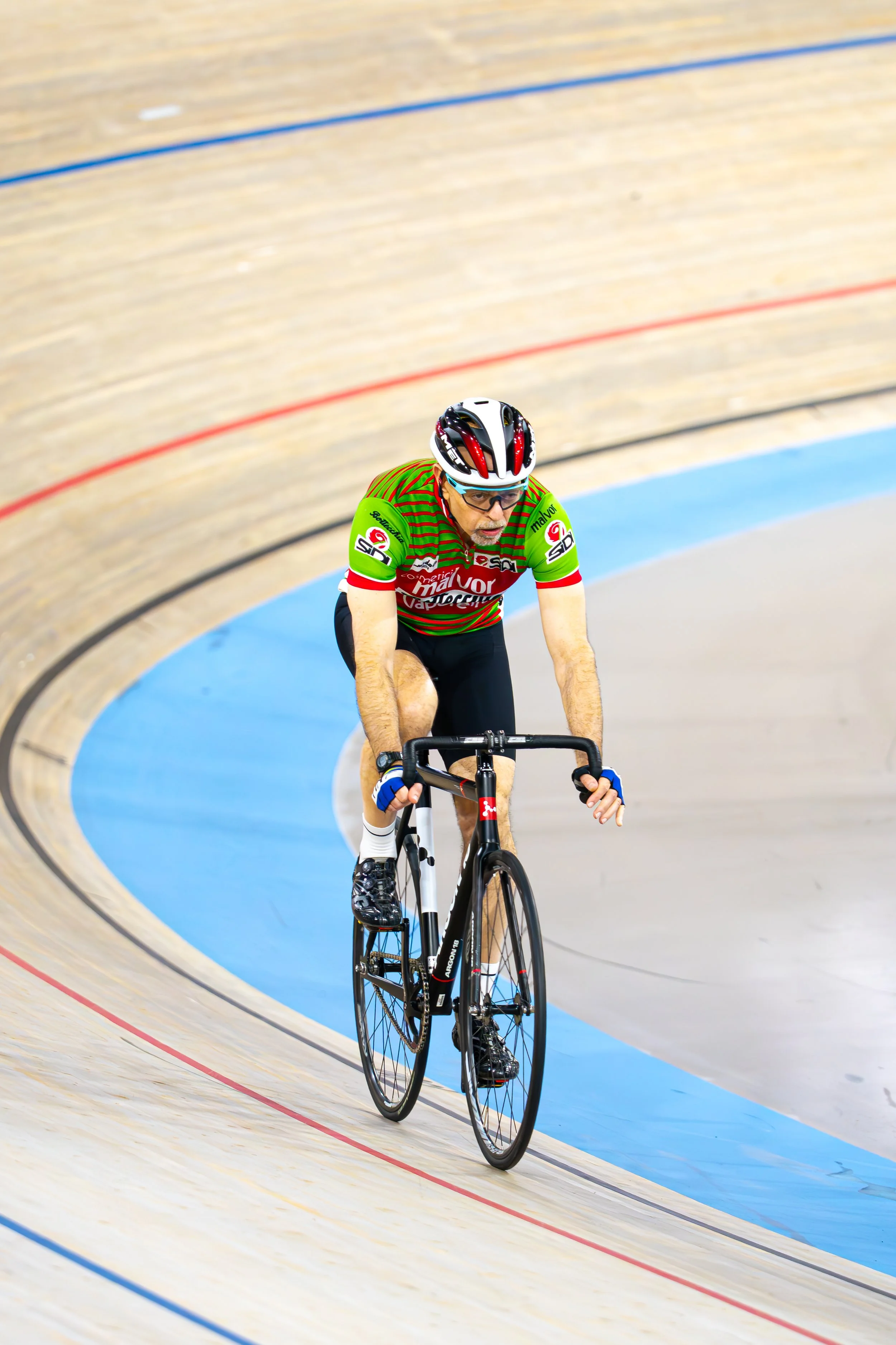 Male cyclist wearing a green and red striped jersey, black shorts, and a white helmet riding on a velodrome track.
