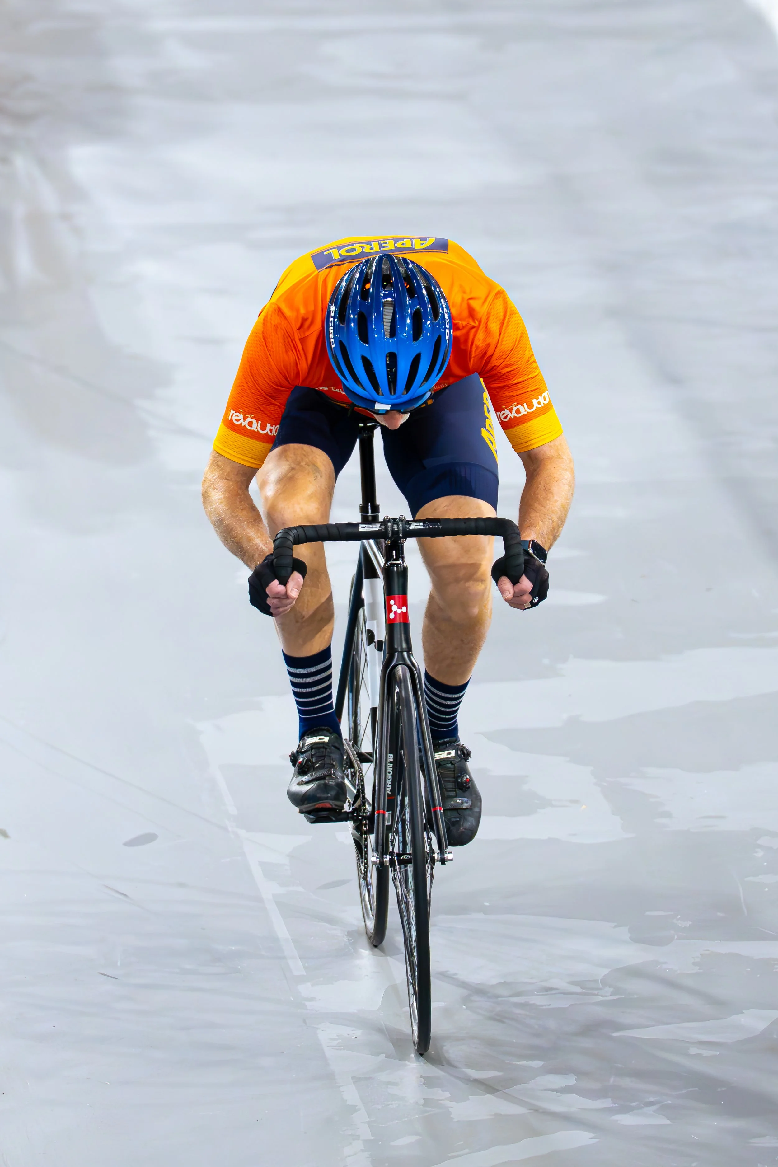 A cyclist wearing an orange jersey, blue helmet, and striped socks rides on a velodrome track.