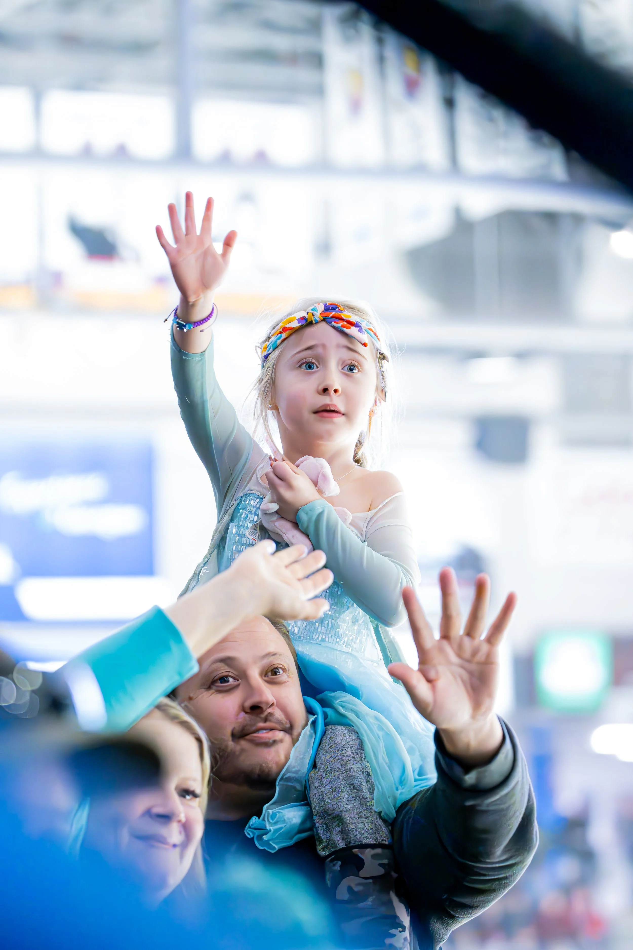 A young girl dressed as a princess with a colorful headband is being carried on the shoulders of a man, likely her father, at an airport. The girl is reaching out with her hand and looks concerned or surprised. The man is smiling and waving while loo