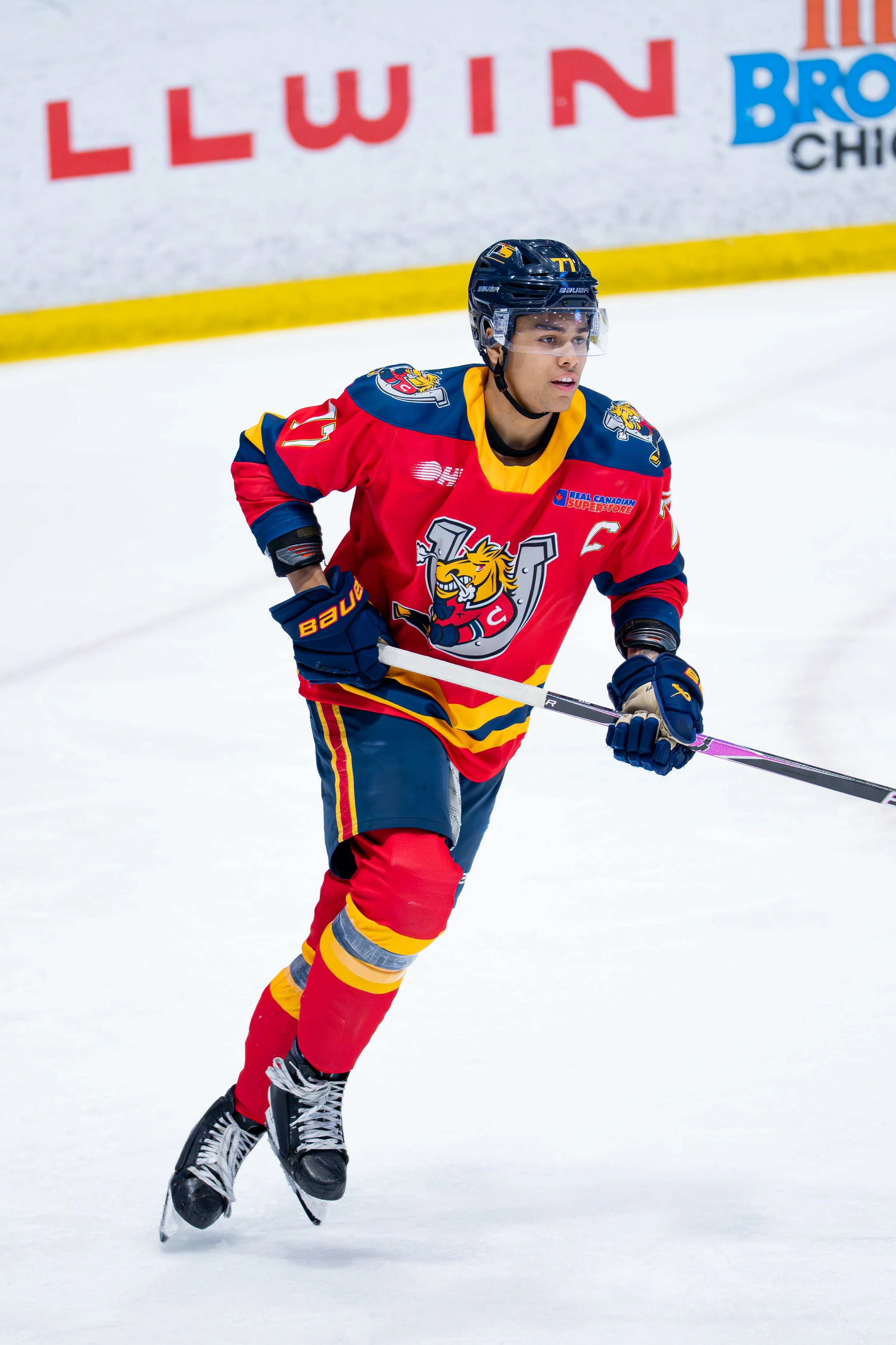Hockey player in red and blue team uniform on ice rink with a focused expression, holding a hockey stick during a game.