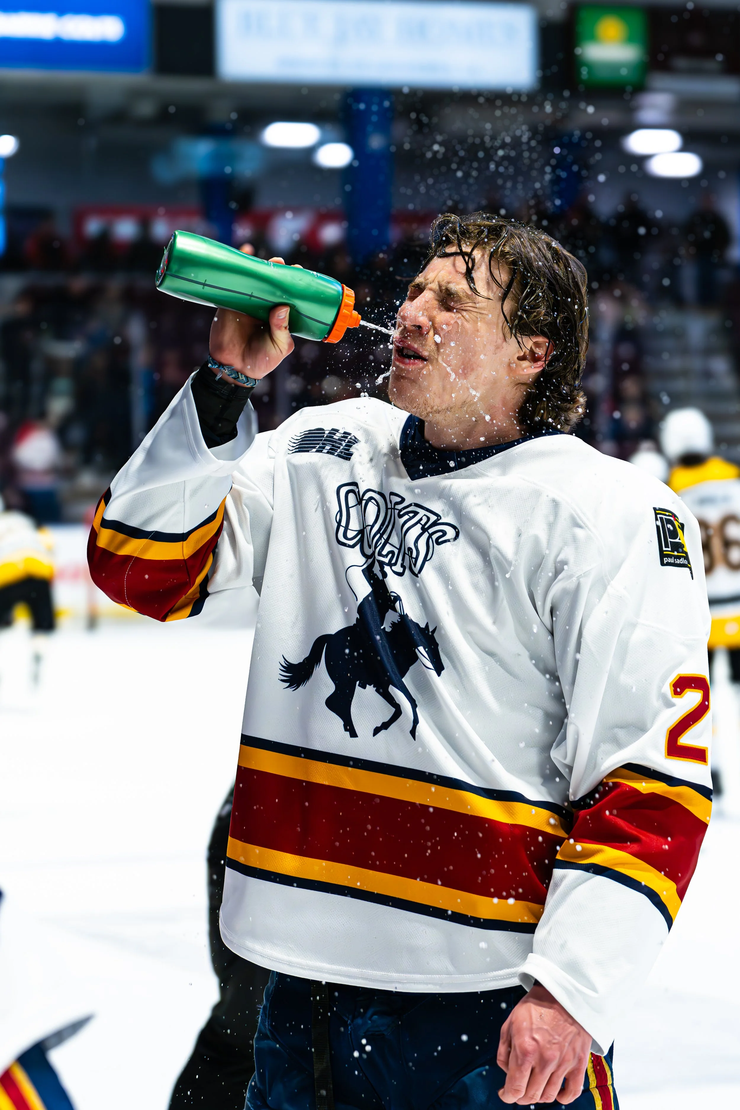 Hockey player in a white jersey drinks water from a green bottle on an ice rink.