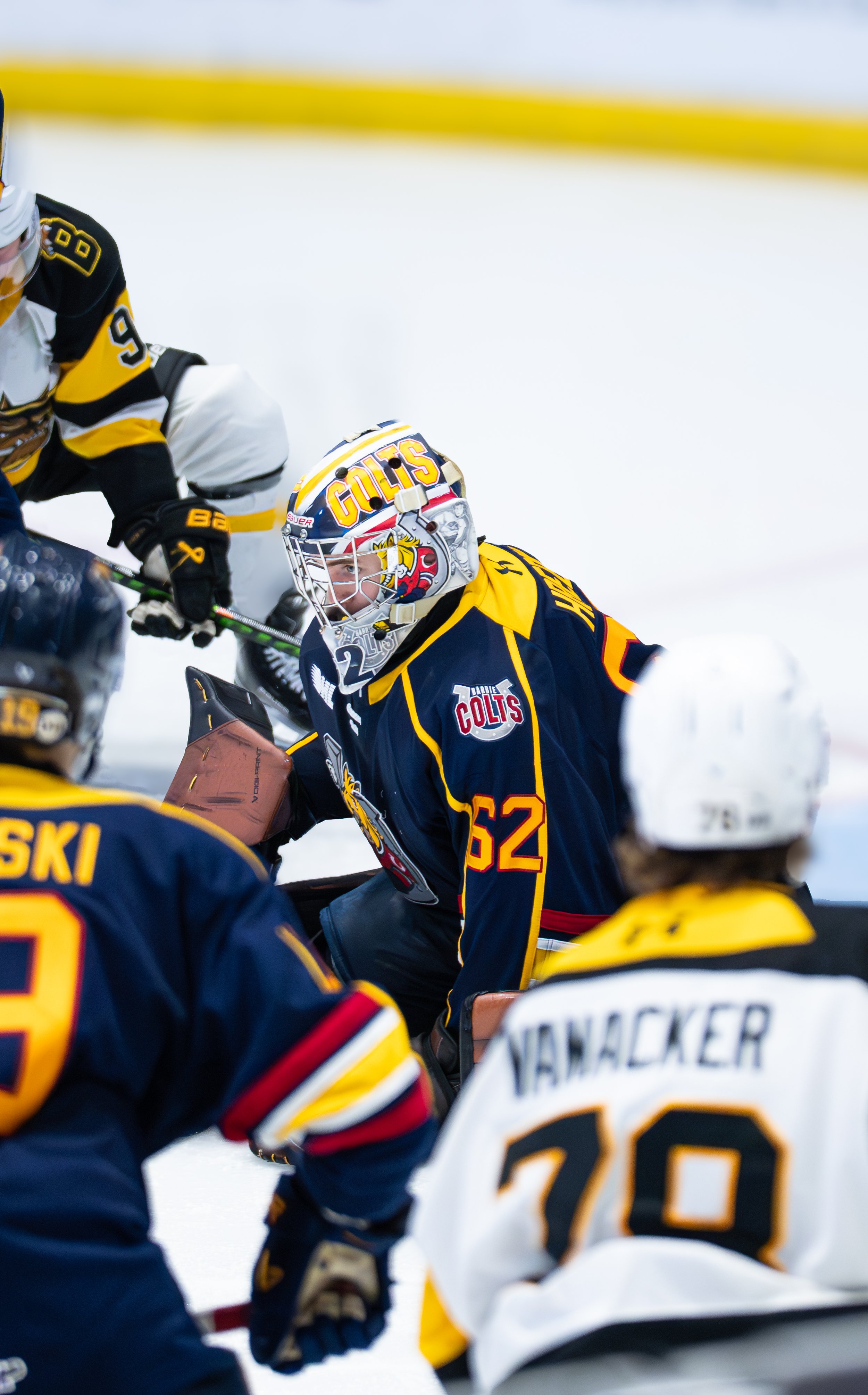 Hockey goalie in a dark blue and yellow uniform with number 52, wearing a helmet with the word 'COLTS' in bold orange letters, crouched in front of the net during a game, surrounded by other players.