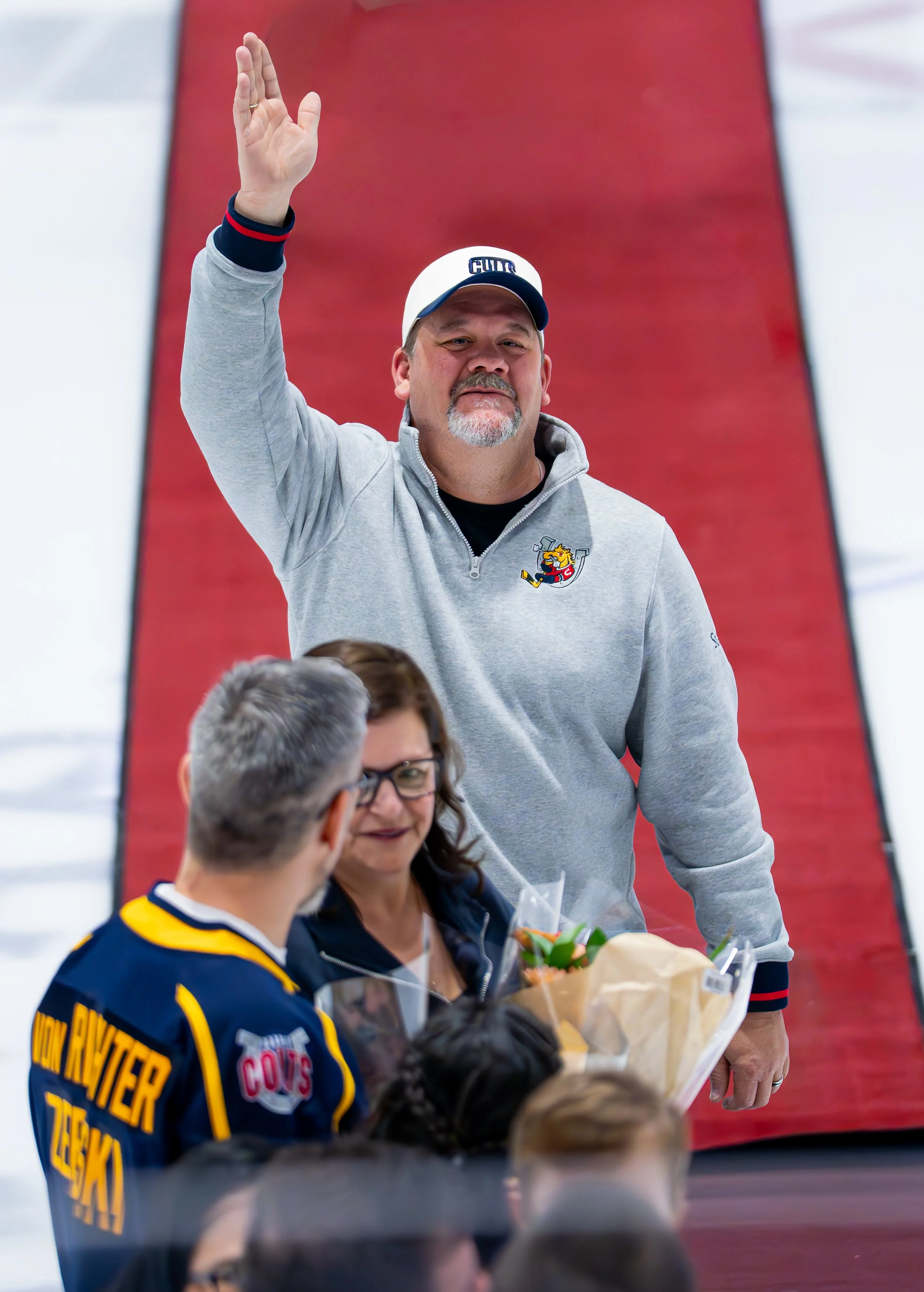 A man in a gray sweatshirt with a hockey logo and a white cap waves to an audience, standing on a red carpet surrounded by people, some holding flowers.