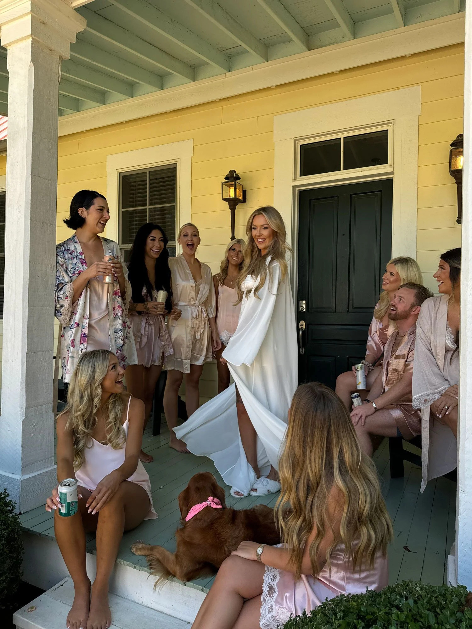 A group of women and men on a porch, celebrating a bride in a white robe. The women are wearing pajamas and holding drinks, with a dog sitting nearby. Everyone is smiling and laughing. Forever After, Instantly Wedding Content Creator in Texas.
