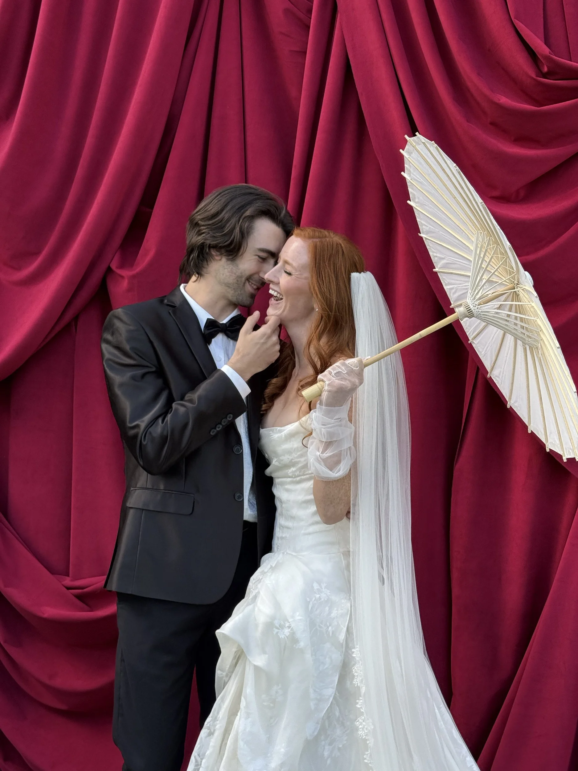 A bride and groom are standing close together in front of a red curtain. The groom is wearing a black tuxedo with a bow tie and has his hand gently touching the bride's face. The bride is dressed in a white wedding gown with lace details, holding a white parasol, and has a veil on her head. They are smiling and appear happy, sharing an intimate moment.