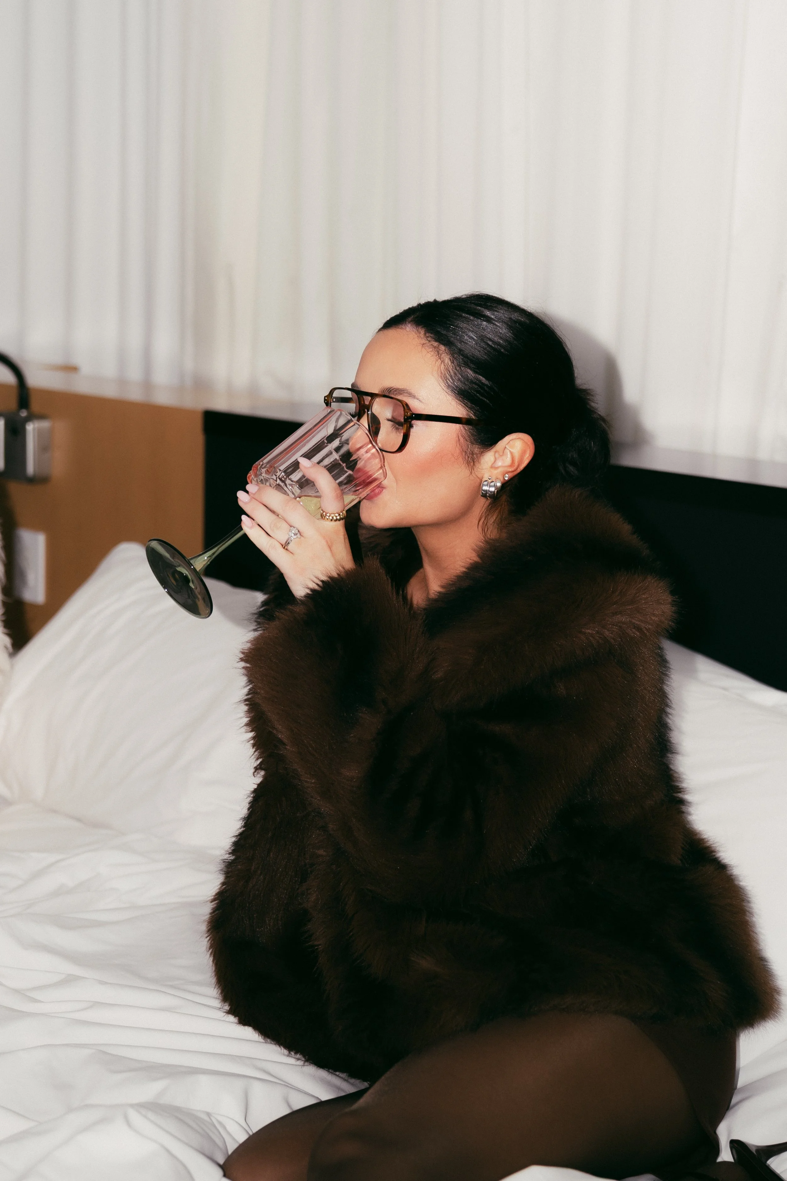 A woman with dark hair and glasses wearing a dark fur coat, drinking from a wine glass, sitting on a bed with white sheets in a hotel room. Kenzie Sikes, Founder of Forever After, Instantly - Wedding Content Creator in TX