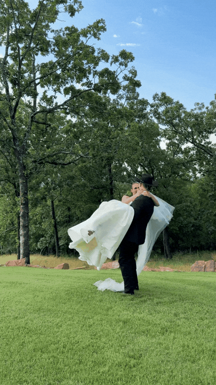 A groom lifting and spinning a bride outdoors on a grassy field with trees and blue sky in the background. Forever After, Instantly Wedding Content Creator in Texas.