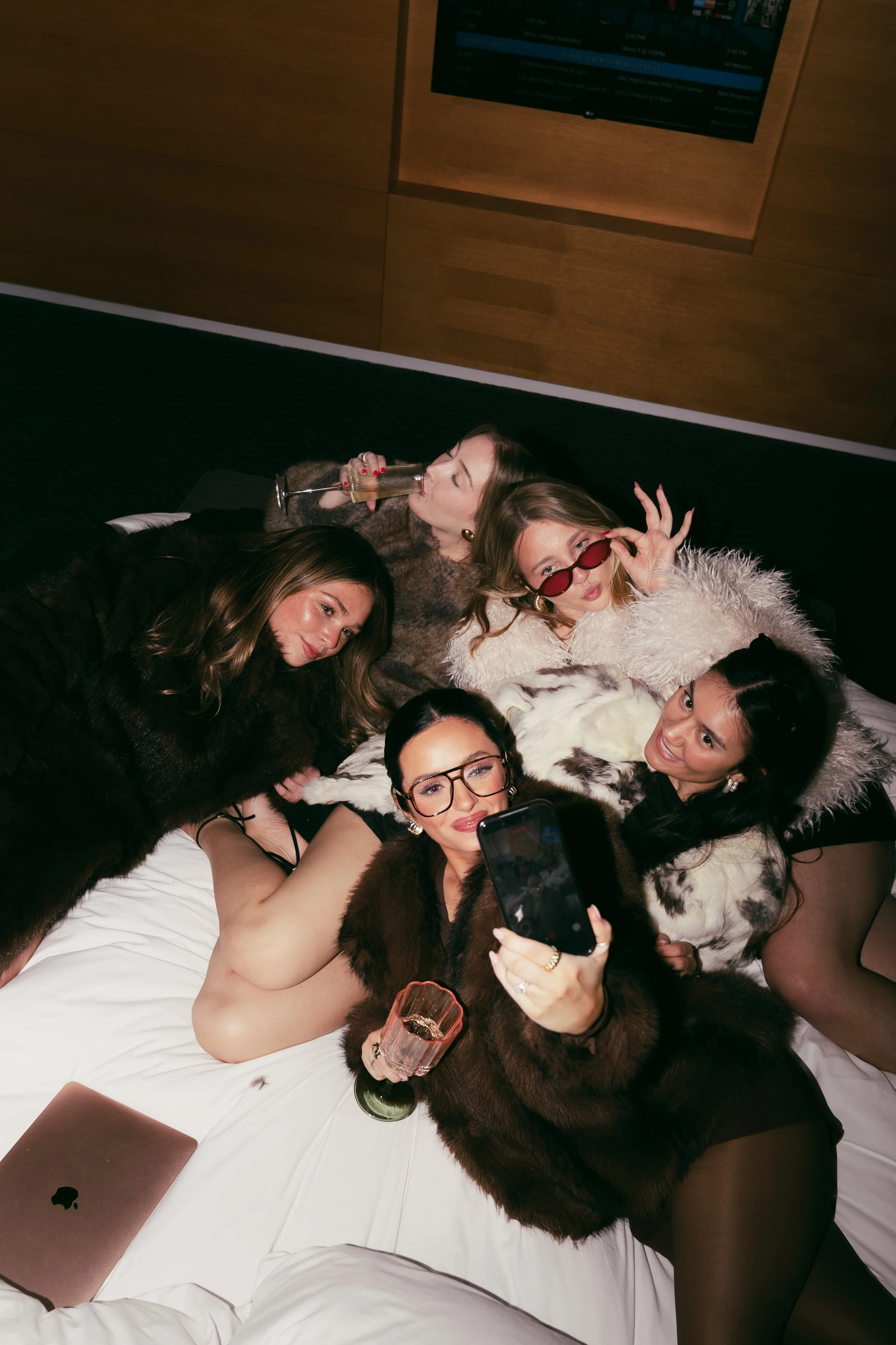 A group of women dressed in glamorous clothing and fur coats are lying on a bed, taking selfies and enjoying drinks. Forever After, Instantly - Wedding Content Creator Team in Texas.