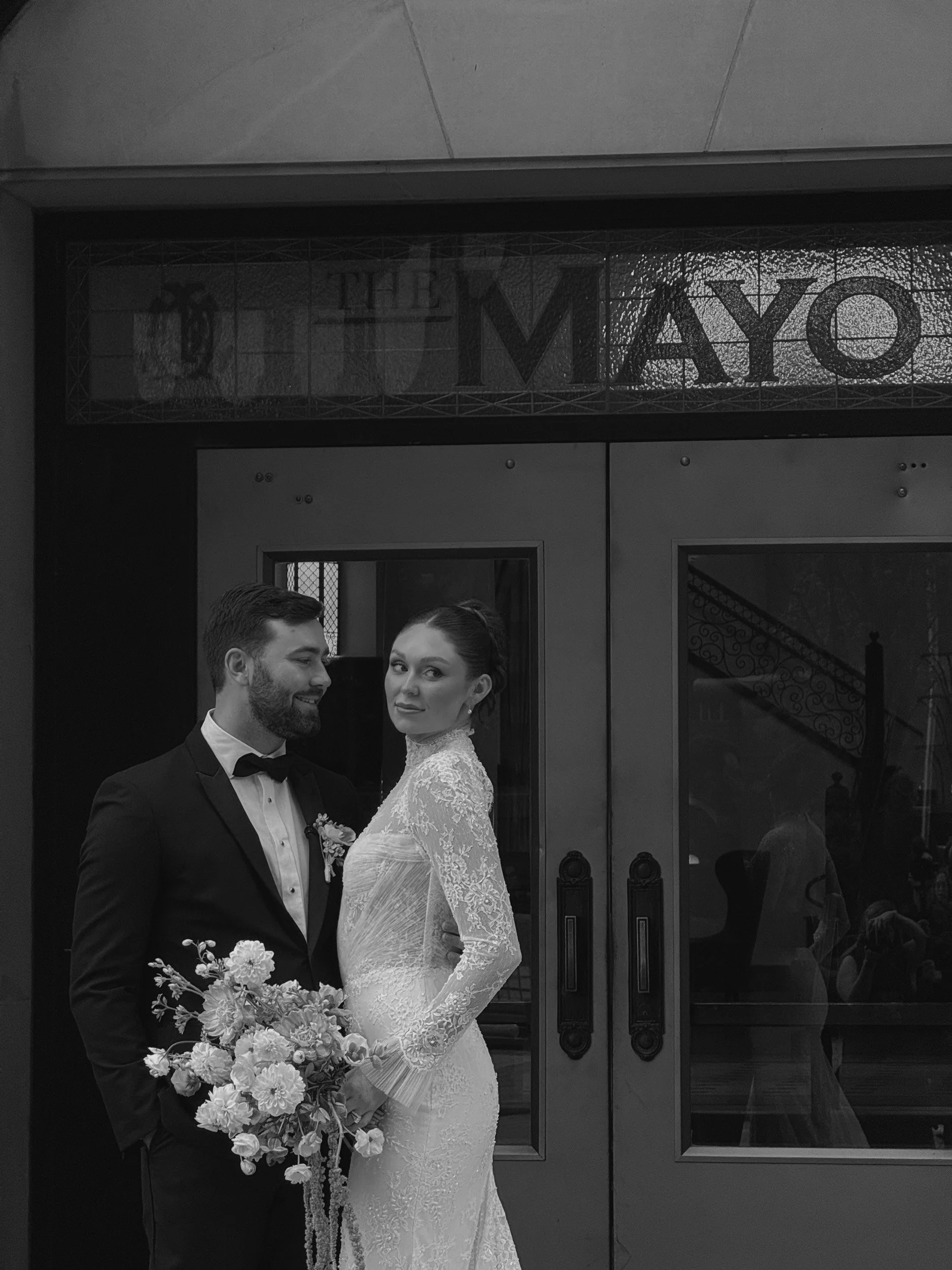 Black and white photo of a bride and groom standing in front of a building. The bride is holding a bouquet of flowers and looking directly at the camera, while the groom is looking at her with a smile.