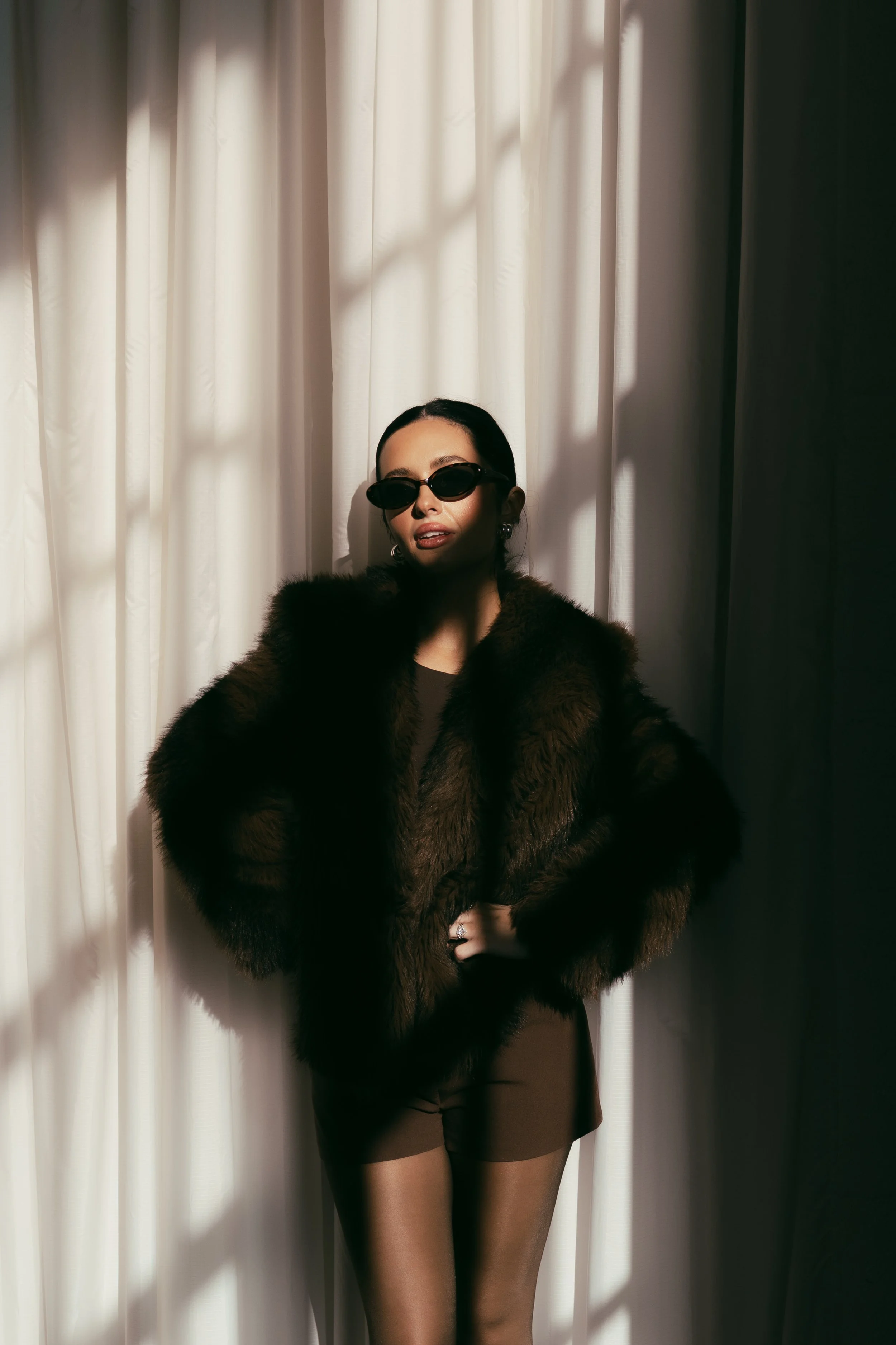 A woman dressed in a brown dress, wearing a fur coat and dark sunglasses, standing in front of a light-colored curtain with shadows cast on it. Kenzie Sikes, Founder of Forever After, Instantly - Wedding Content Creator