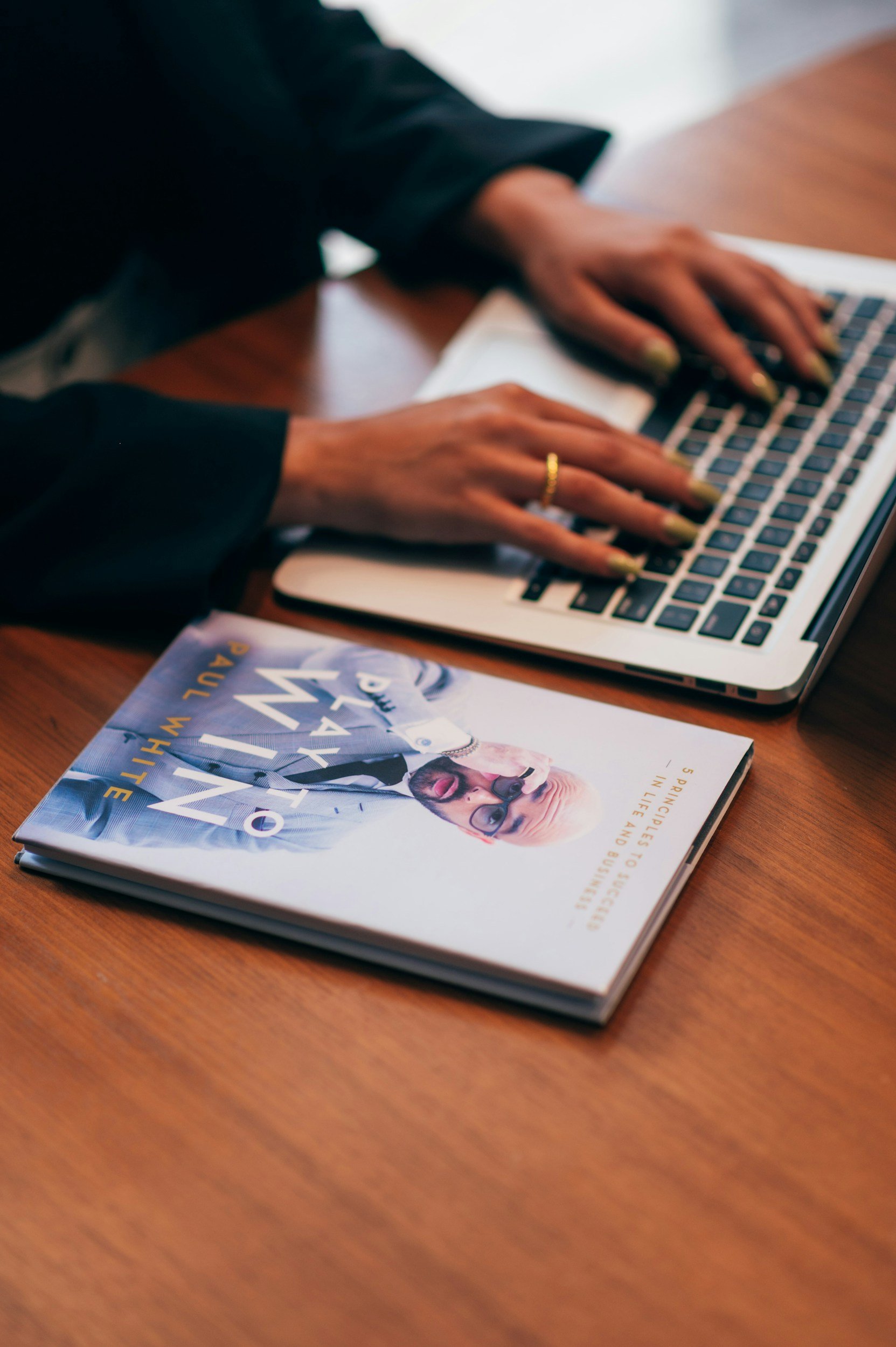A person typing on a laptop at a wooden desk with a magazine featuring a man wearing sunglasses.