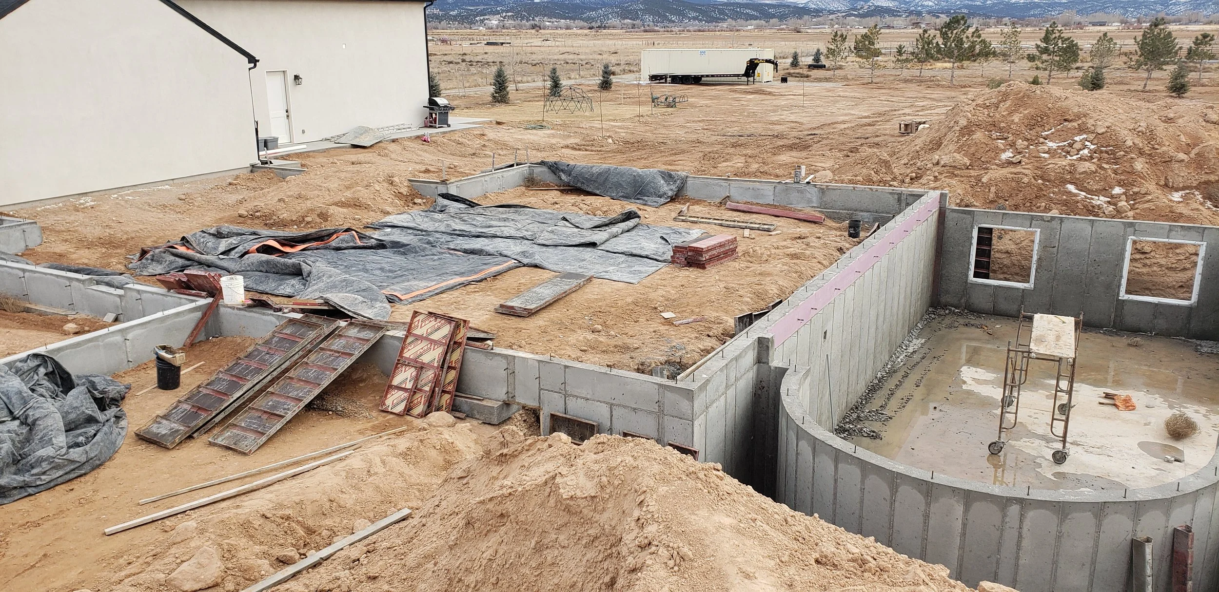 Construction site of a house foundation with concrete walls and an unfinished basement, surrounded by dirt and construction materials, under an overcast sky.