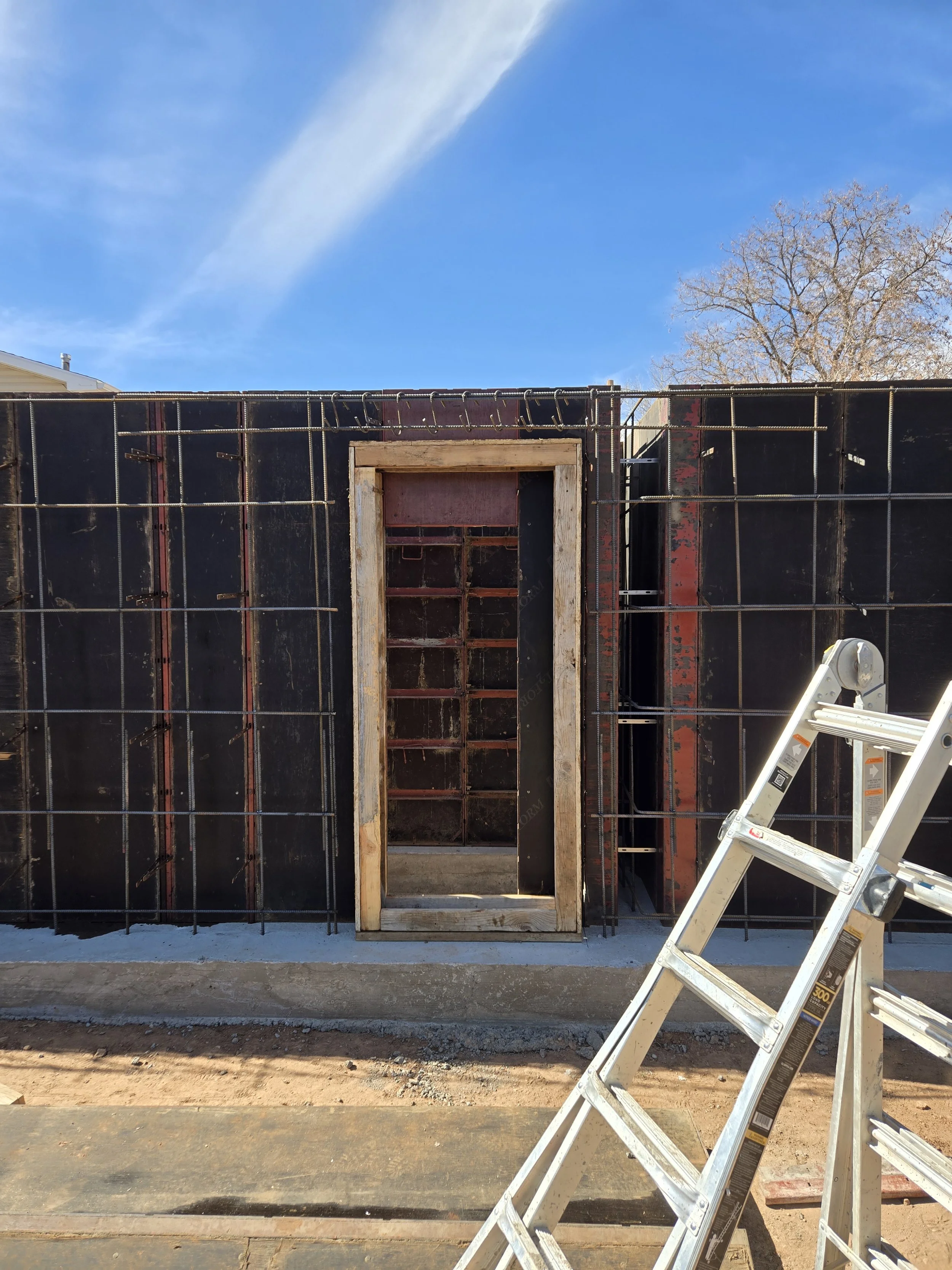 Construction site with a partially built wall made of black sheathing and wooden framing. A metal ladder is leaning against the wall, and a cloudy blue sky is visible in the background.