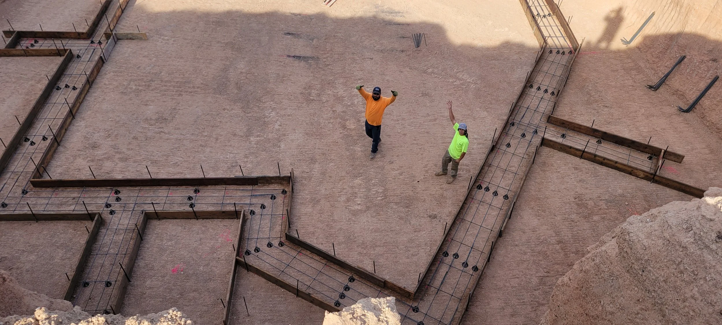 Two construction workers, one in an orange shirt and the other in a neon green shirt, standing inside a construction site with rebar framework for a foundation or structure.