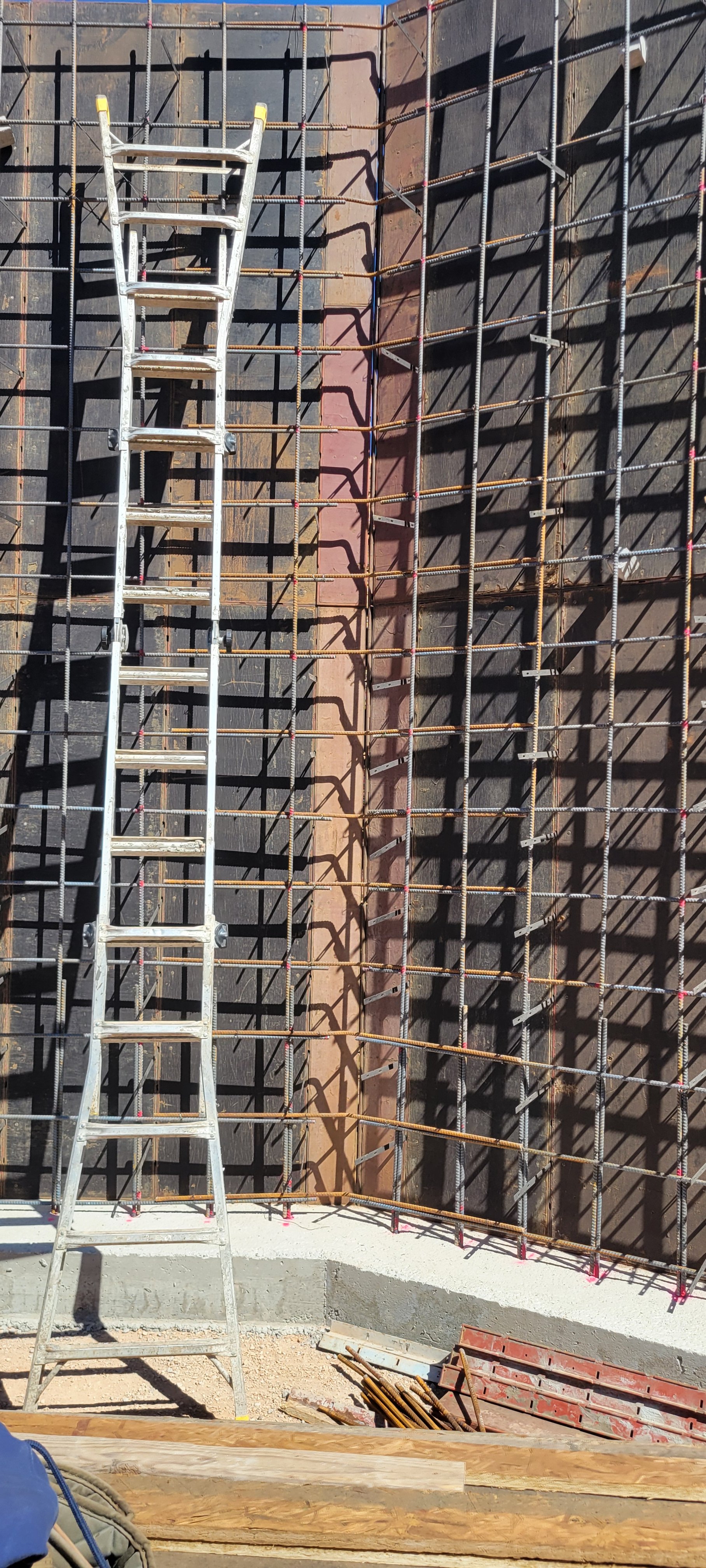 Construction site with metal rebar grid being prepared for concrete, a measuring ladder leaning against the rebar, and construction materials on the ground.