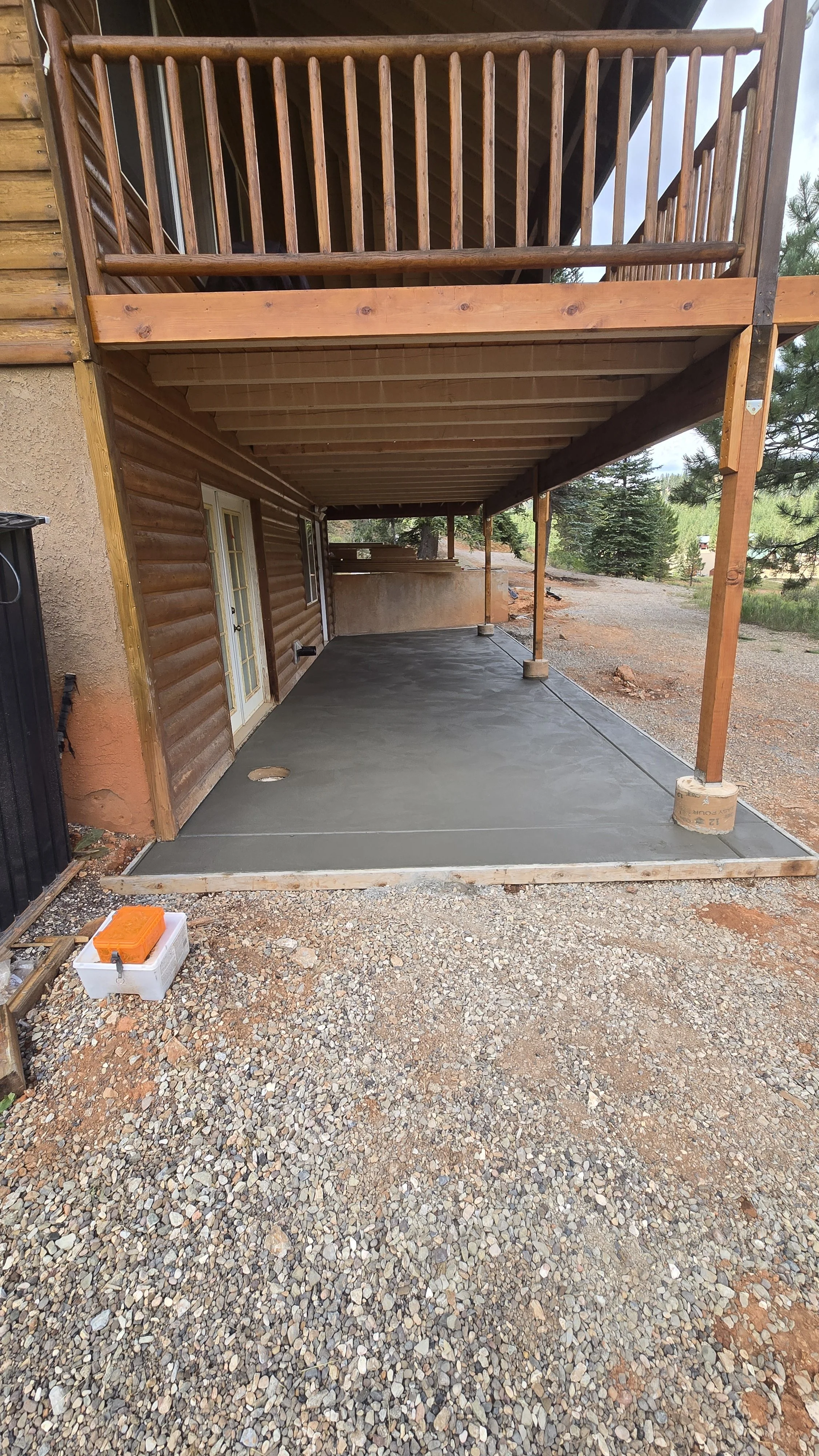 Finished concrete patio beneath a wooden deck with a gravel surrounding area, in a backyard with trees.