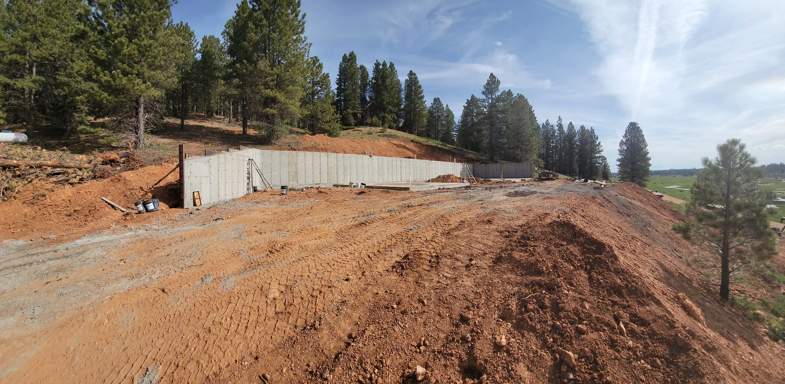 Construction site on a hillside with concrete retaining wall, trees, and dirt in the foreground.