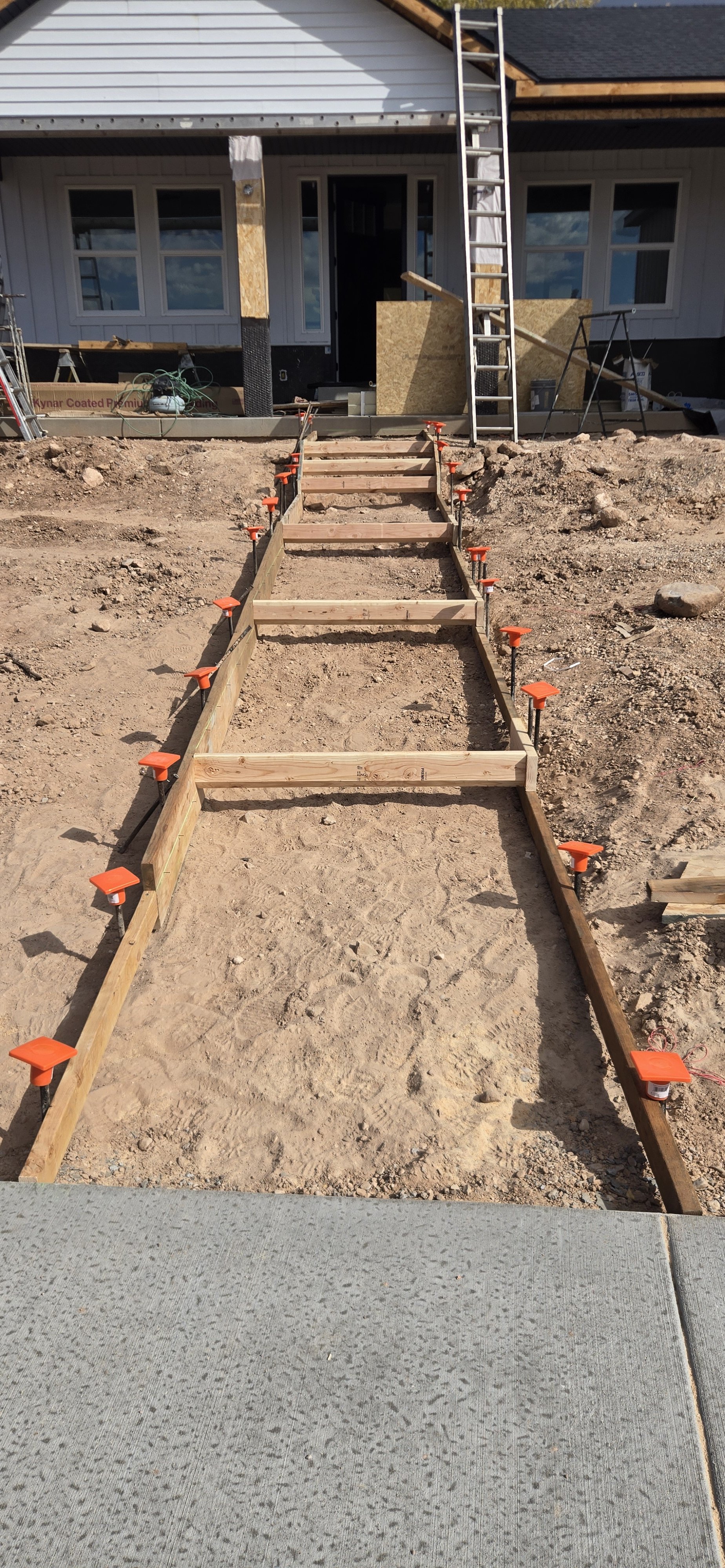 Construction site showing a wooden stairway under construction, with the framing and supports visible, leading up to a house with scaffolding and construction materials.