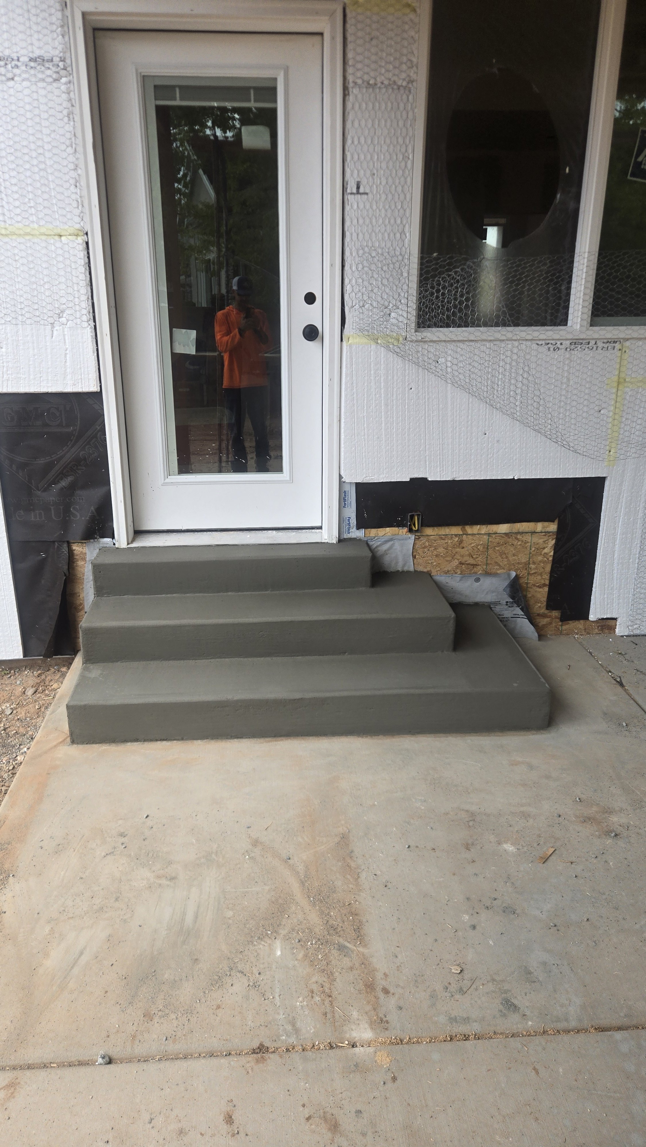 Newly constructed front porch with three gray concrete steps leading to a white door with a glass panel. The area around the porch shows signs of ongoing construction, with exposed insulation and unfinished surfaces.
