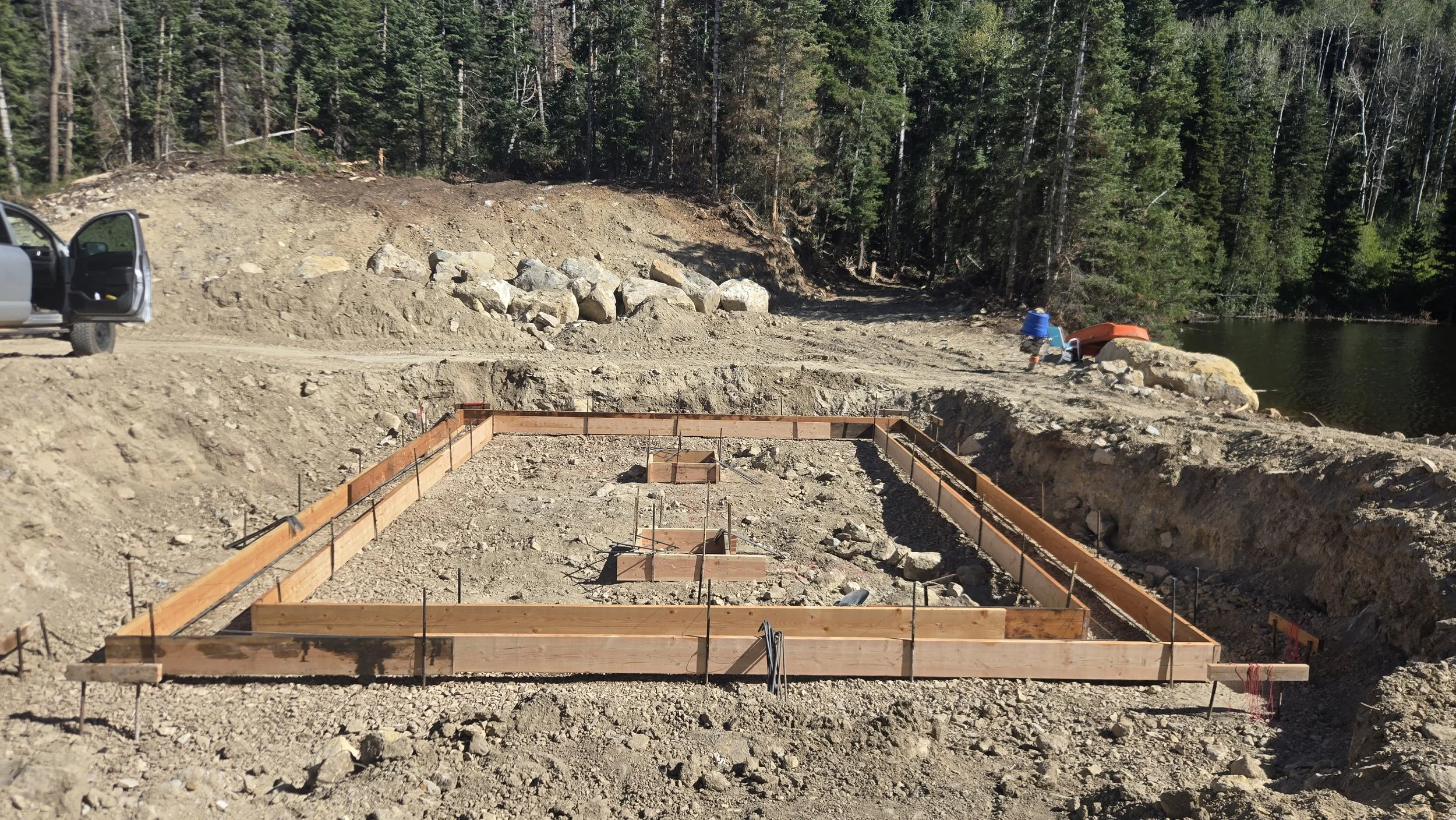Construction site in a forested area with wooden framing for concrete foundation, dirt, and rocks, near a body of water with trees in the background.