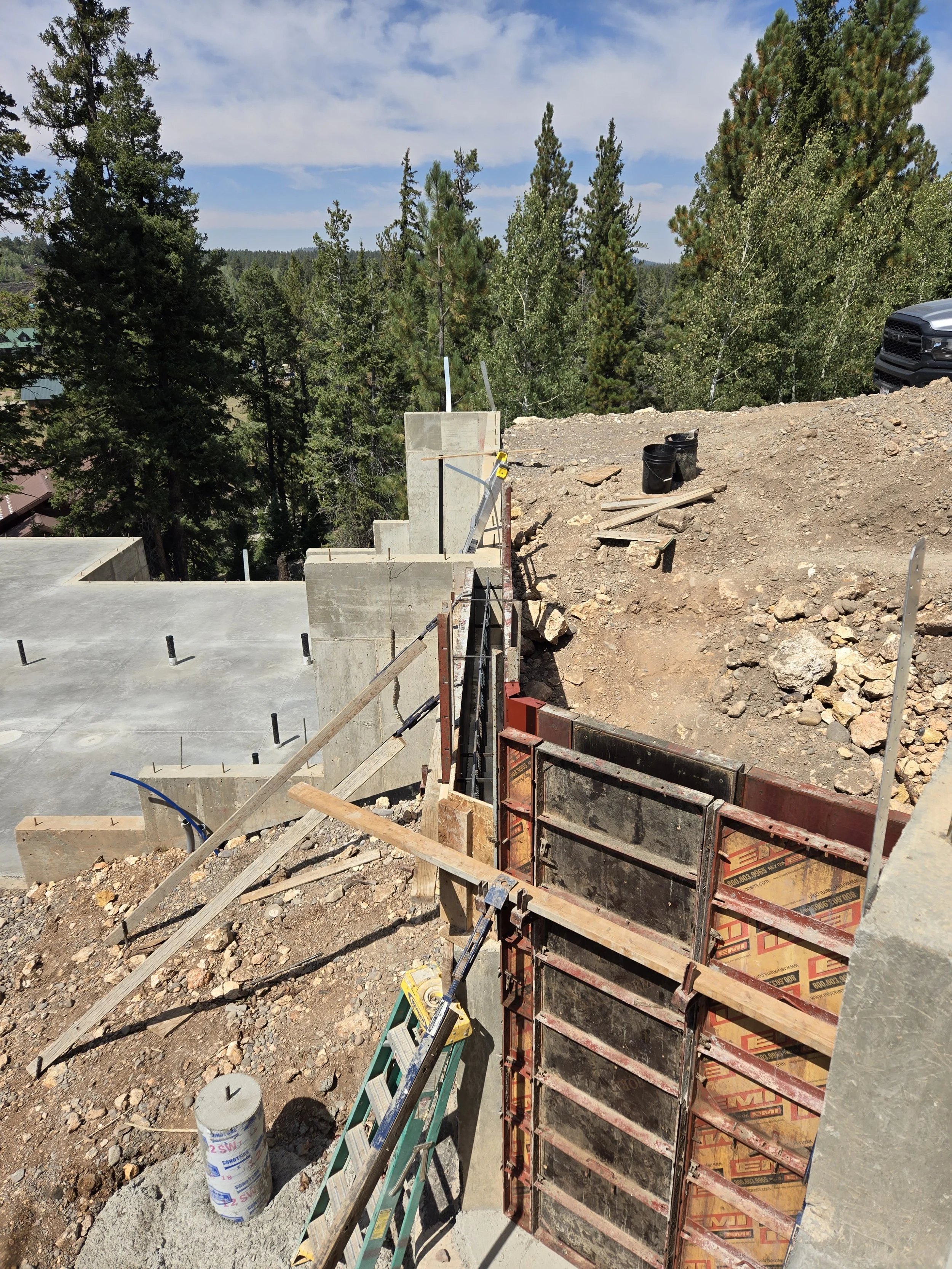Construction site with a concrete foundation and framing for a building, surrounded by trees and hills under a partly cloudy sky.
