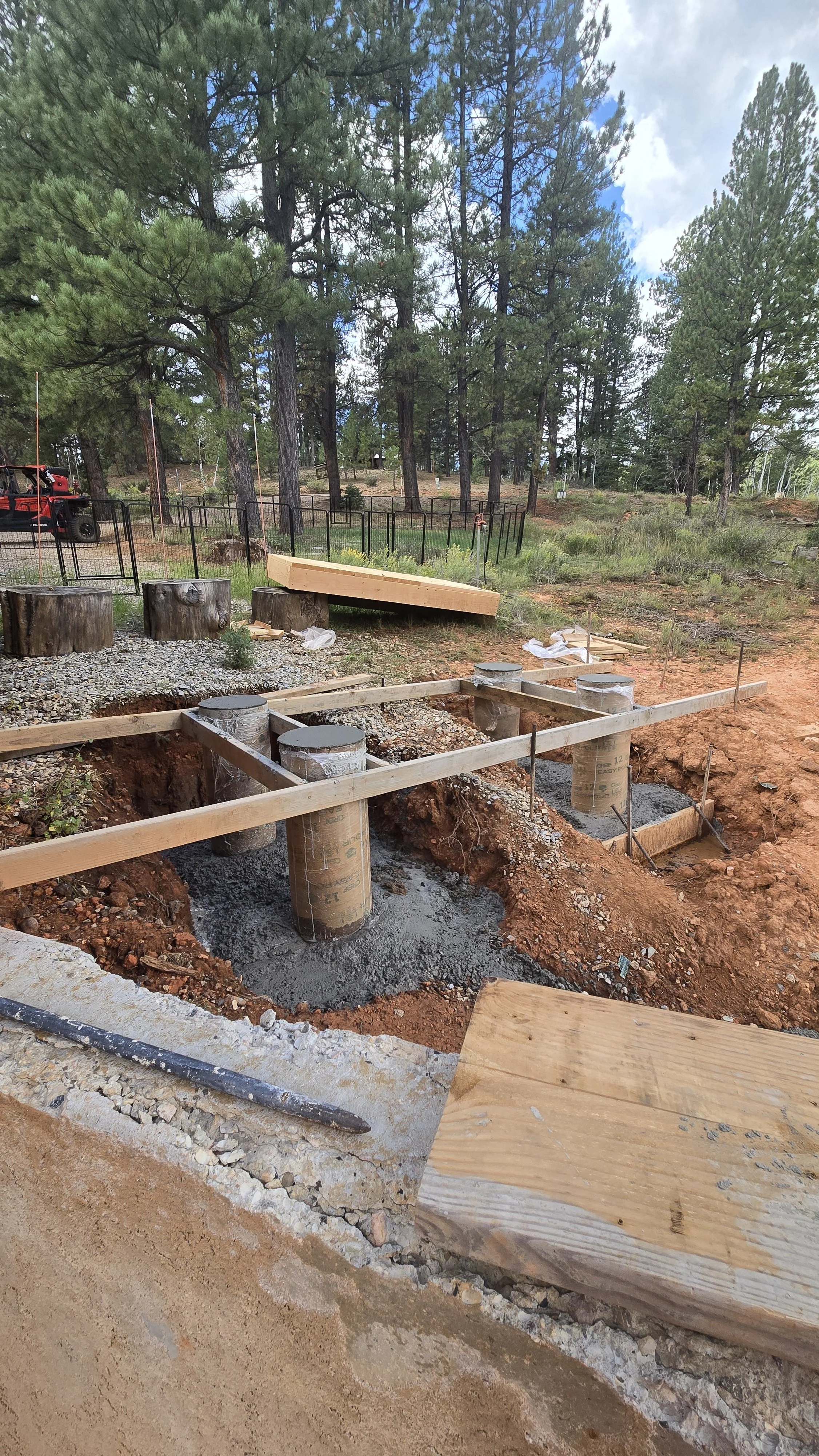 Construction site in a forested area with concrete pillars and wooden forms for foundation work.