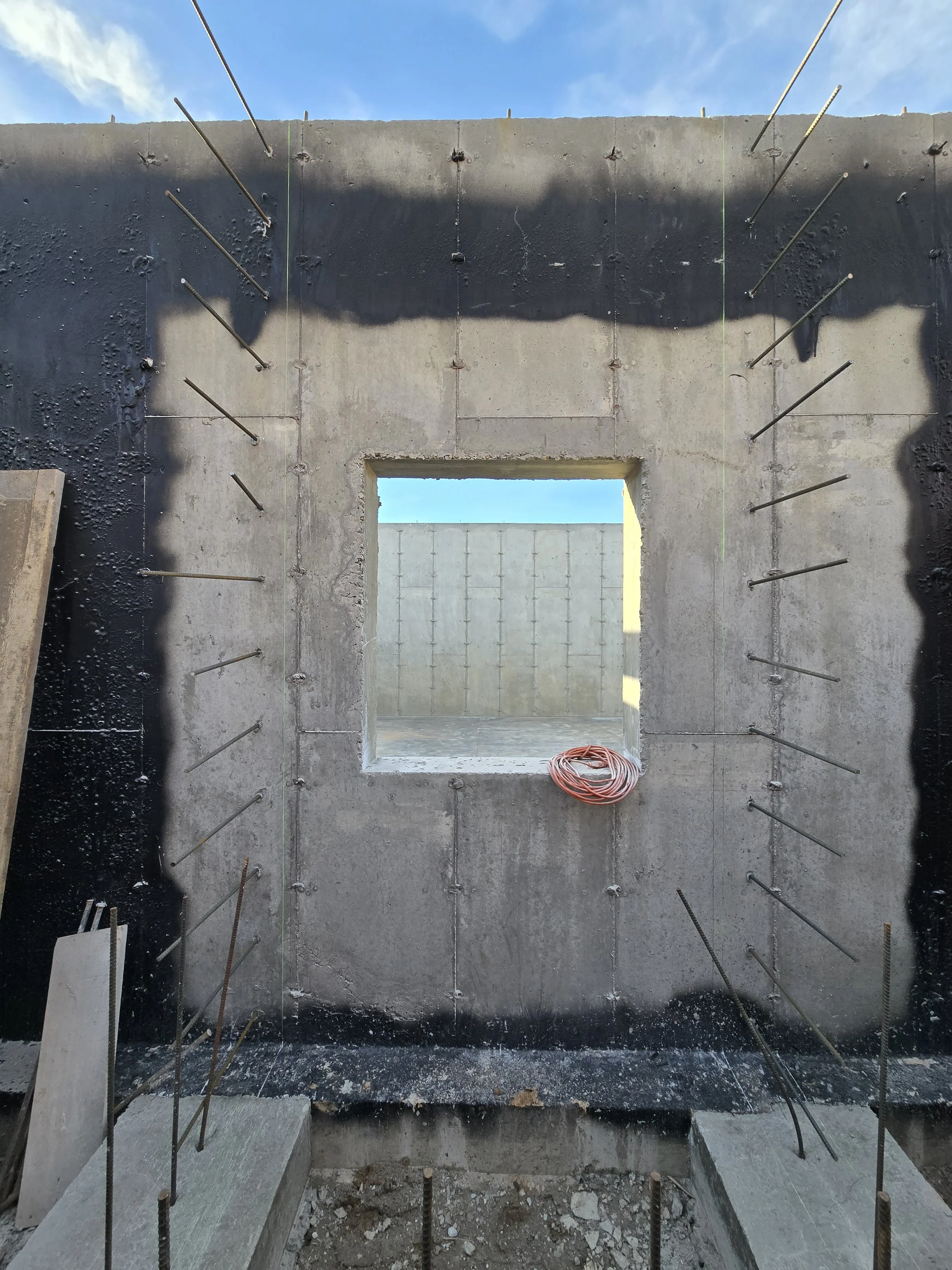 A concrete wall with protruding metal rebar rods and a rectangular opening cut out, with construction materials and a coiled cable visible at the bottom, under a blue sky with some clouds.