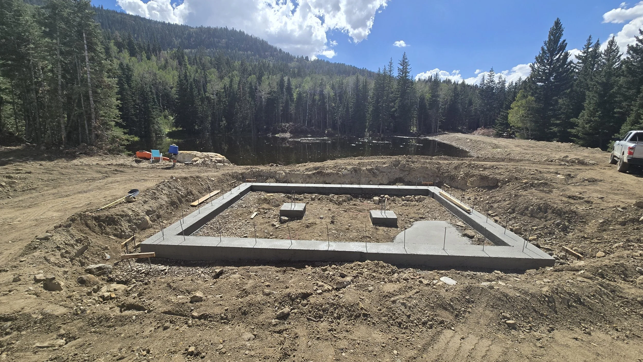 Construction site with a foundation in a forested mountain area, with tools and vehicles nearby, lake in the background, and partly cloudy sky.