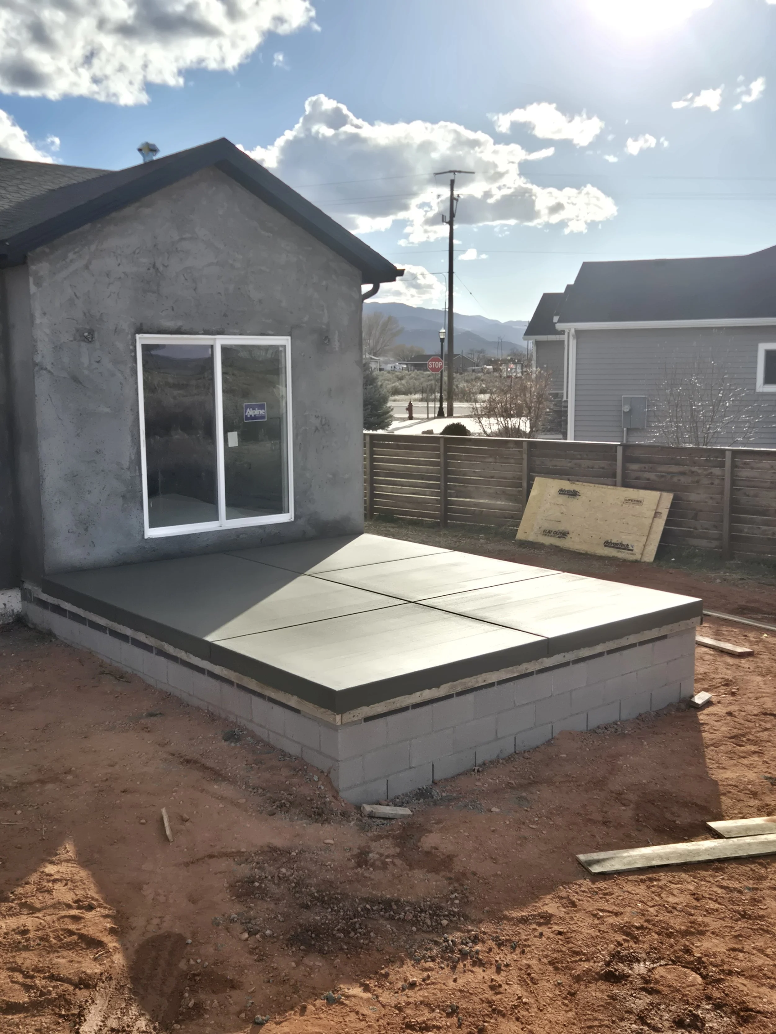 A construction site with a partially built small house, concrete patio slab, and a wooden fence. The house has a window and exterior wall showing a sign that says 'Alpine'. There is a clear sky with some clouds, and mountains can be seen in the backg
