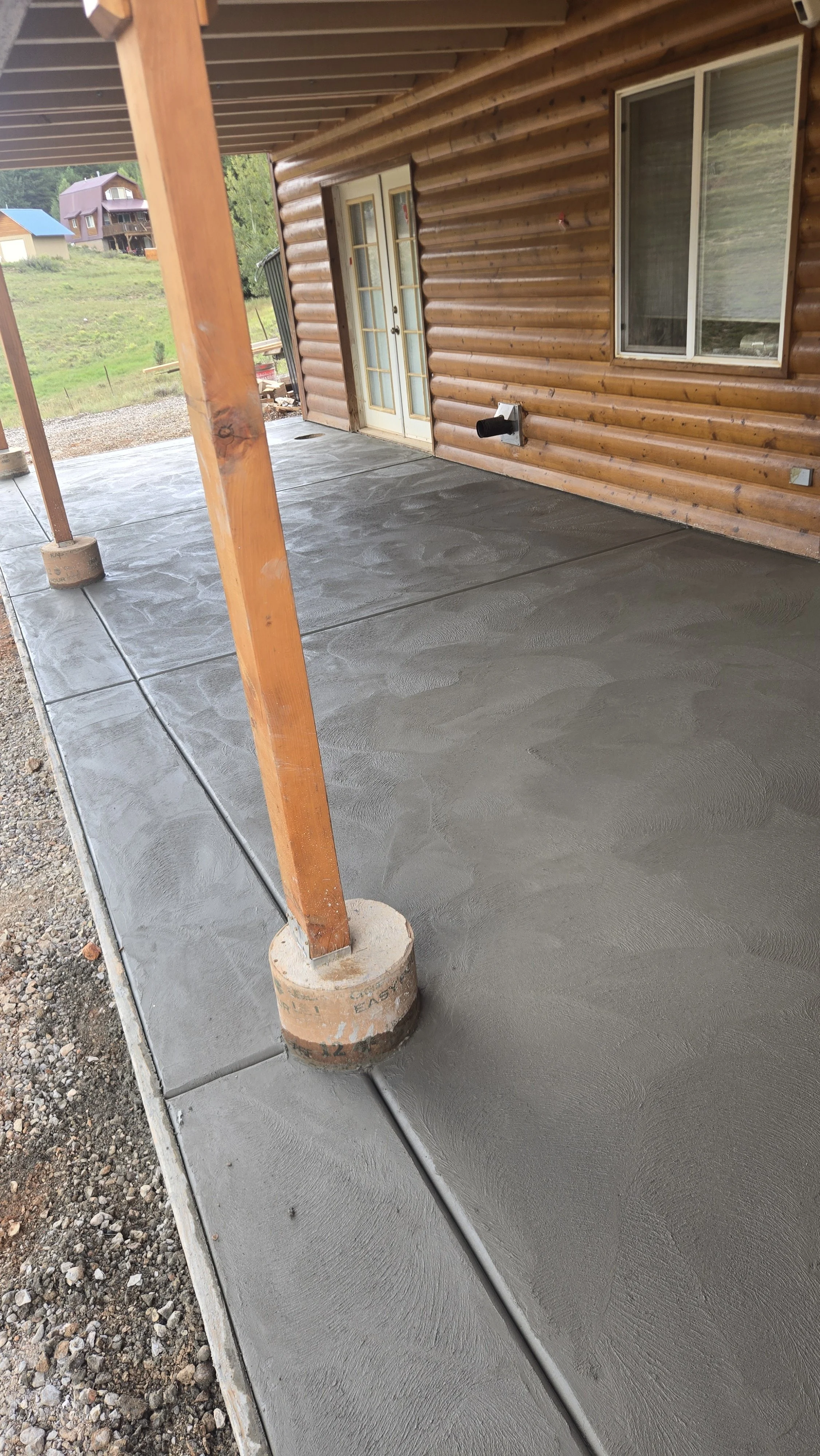 Newly poured concrete on a porch with wooden support beams and a log cabin exterior wall with two windows and a door