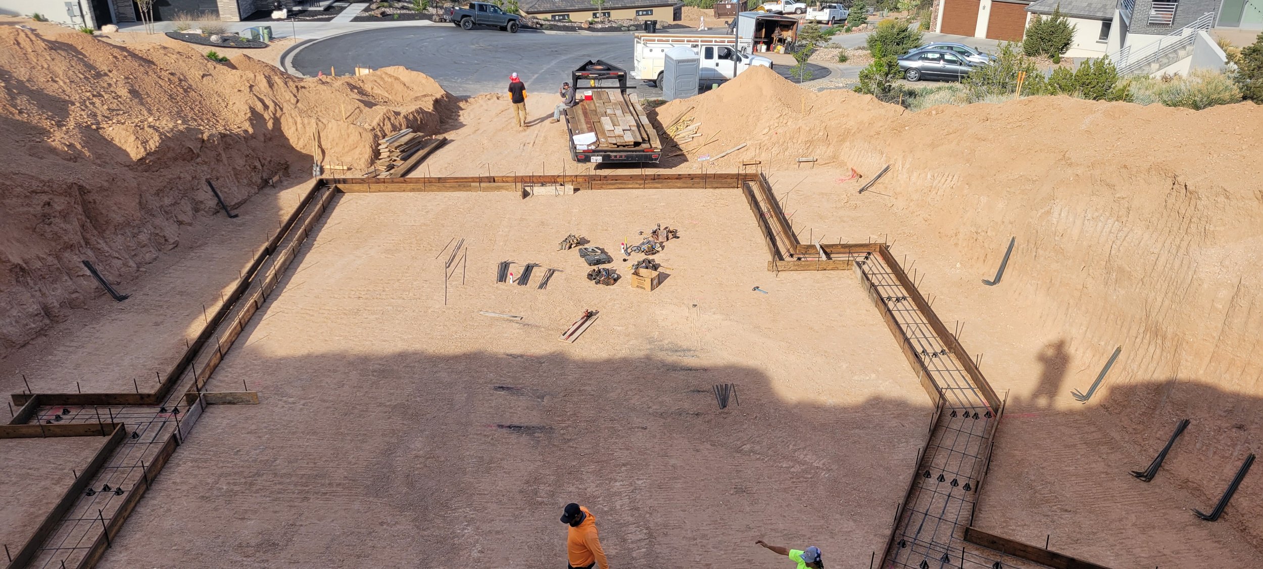 Construction site with excavated foundation, steel rebar framework, and workers preparing the area for concrete pouring in a residential neighborhood.