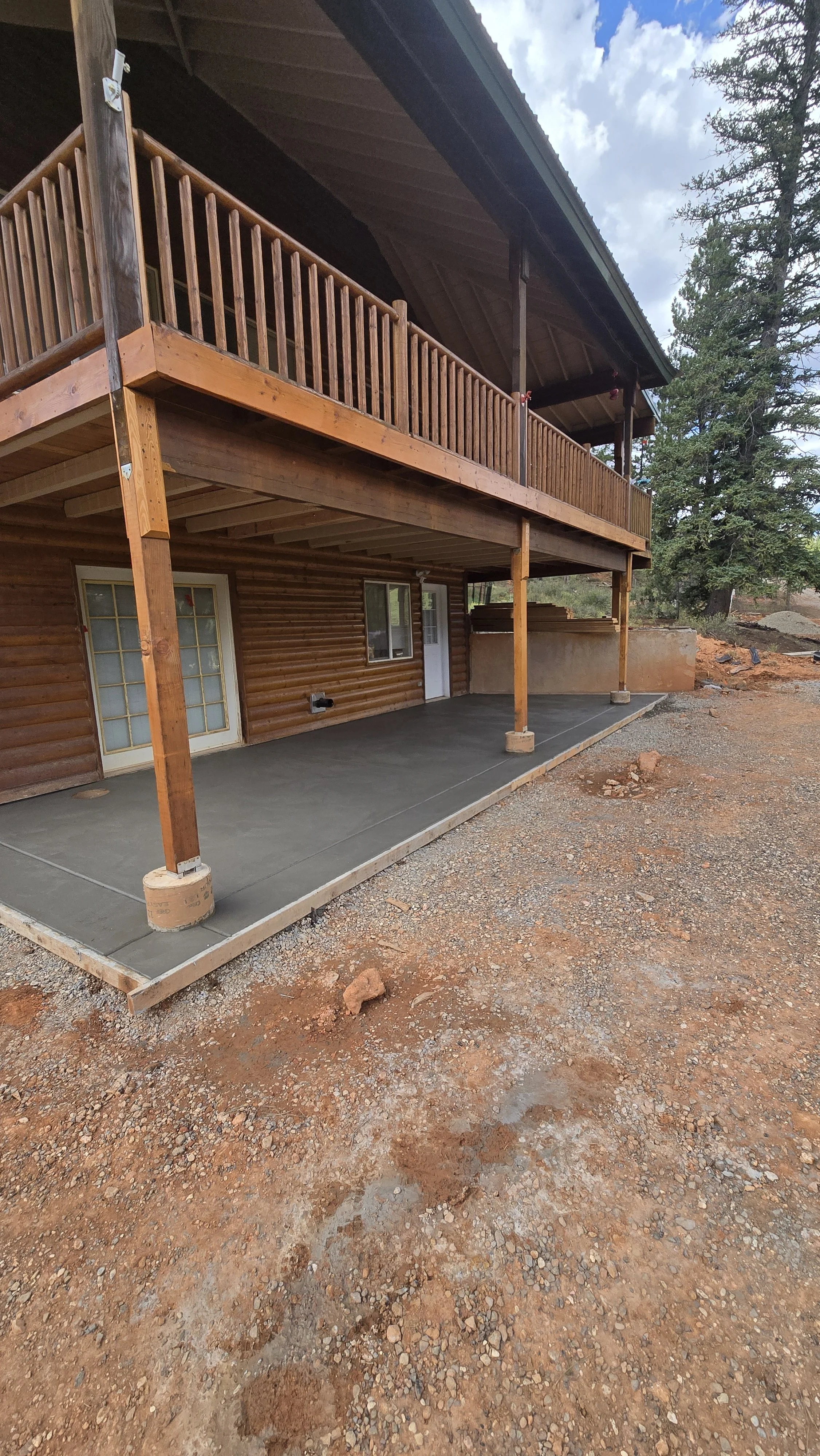 Exterior view of a two-story house with a wooden balcony on the upper level, supported by wooden posts. The ground level has a concrete patio and some landscaping, with a dirt and gravel area in the foreground, surrounded by trees.