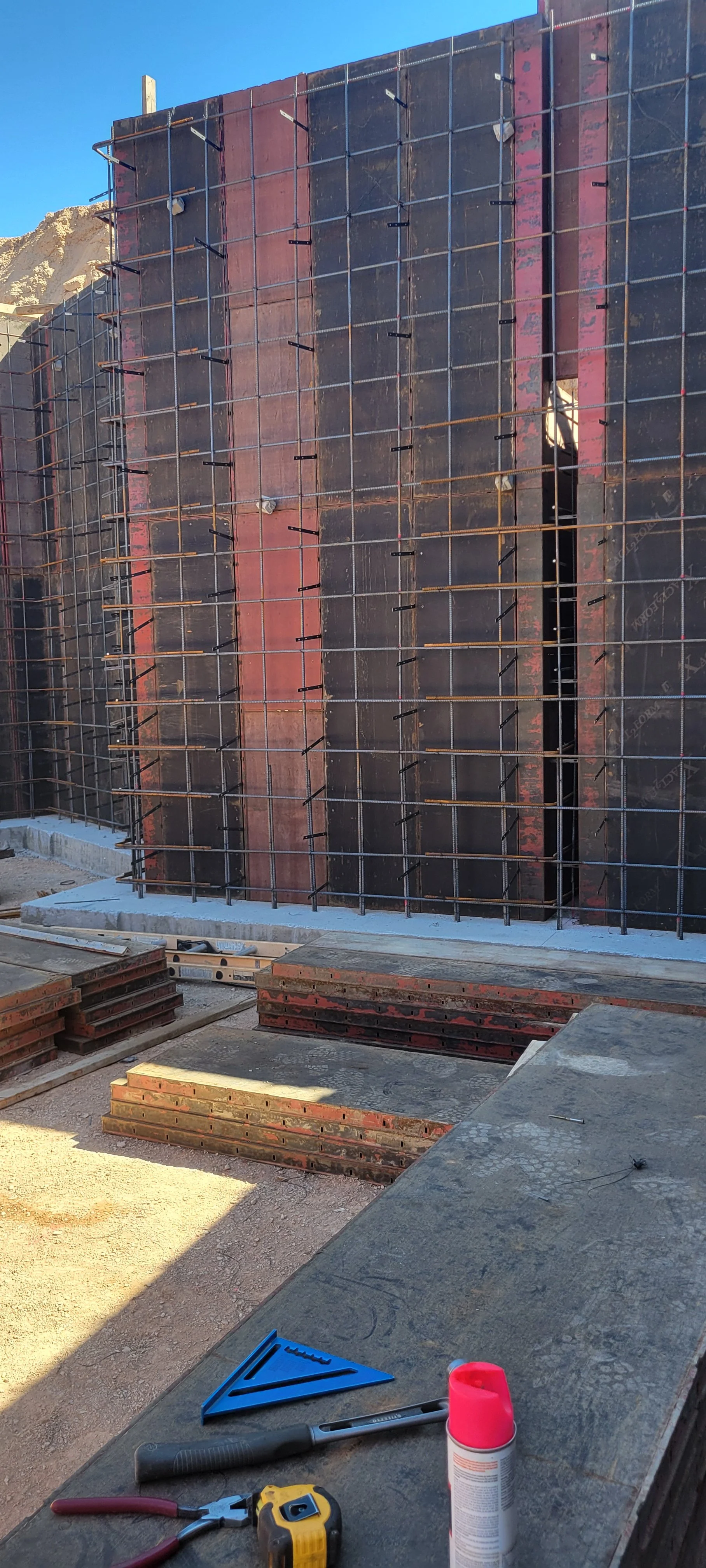 Construction site with a concrete wall under construction reinforced with steel rebar and wooden planks, construction tools and materials on the foreground, clear blue sky in the background.