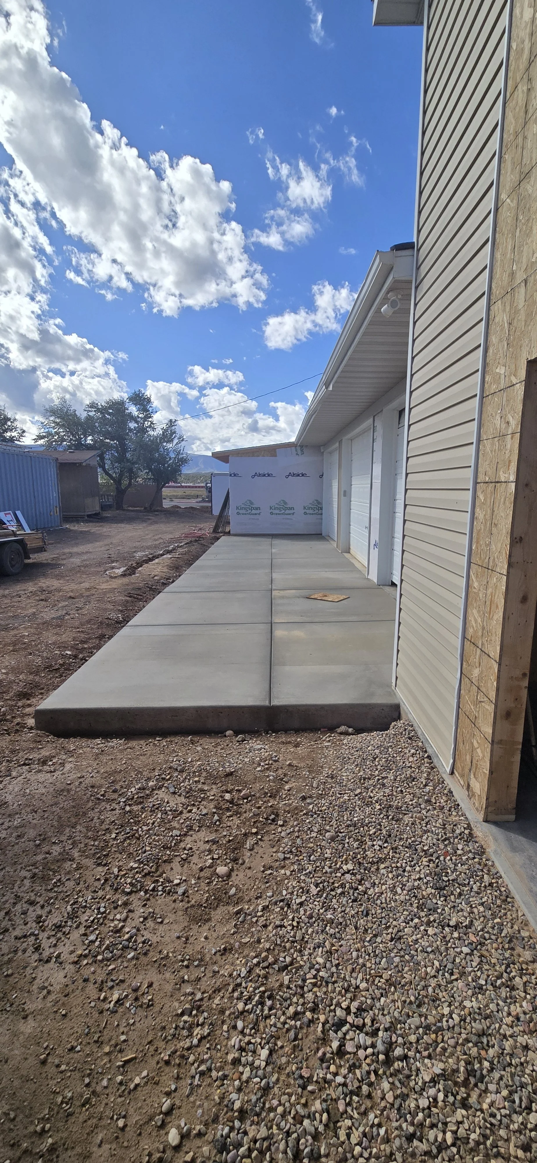 Newly poured concrete sidewalk adjacent to a building with beige siding, under a partly cloudy sky.