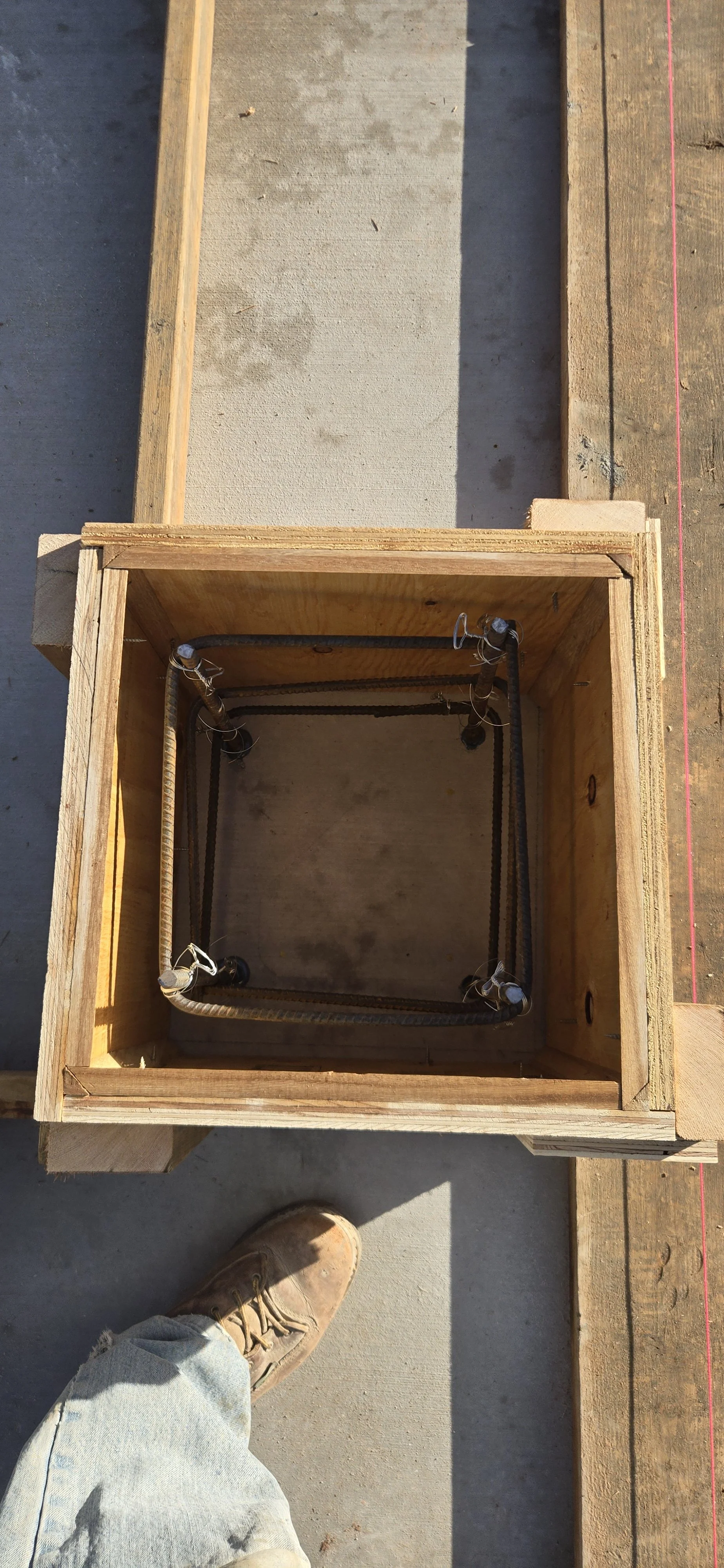 View from above showing a wooden form with rebar inside for concrete reinforcement, likely part of a construction project, on a concrete floor with a shadow cast.