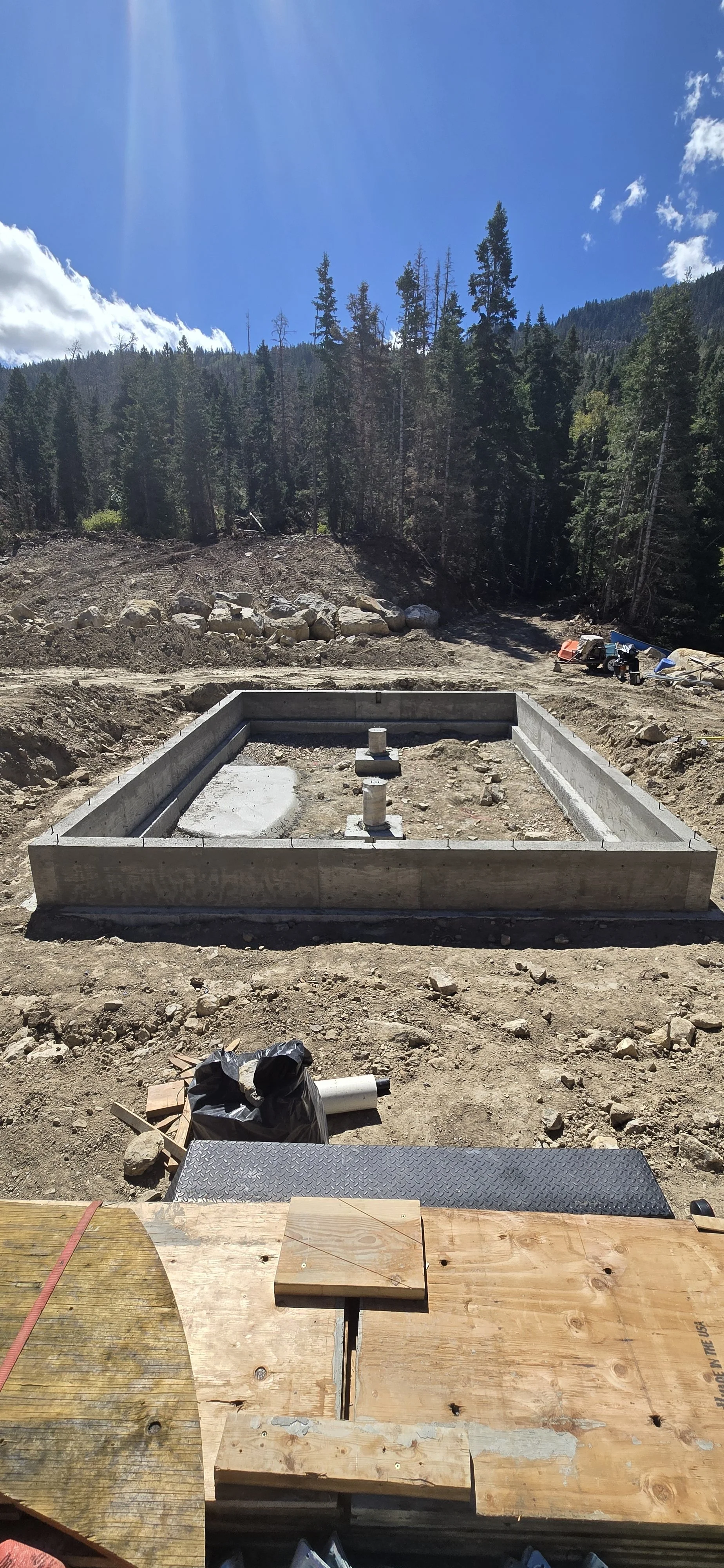 Construction site in a mountainous forest area with a concrete foundation for a building and pipes protruding, surrounded by dirt and construction materials under a blue sky with clouds.