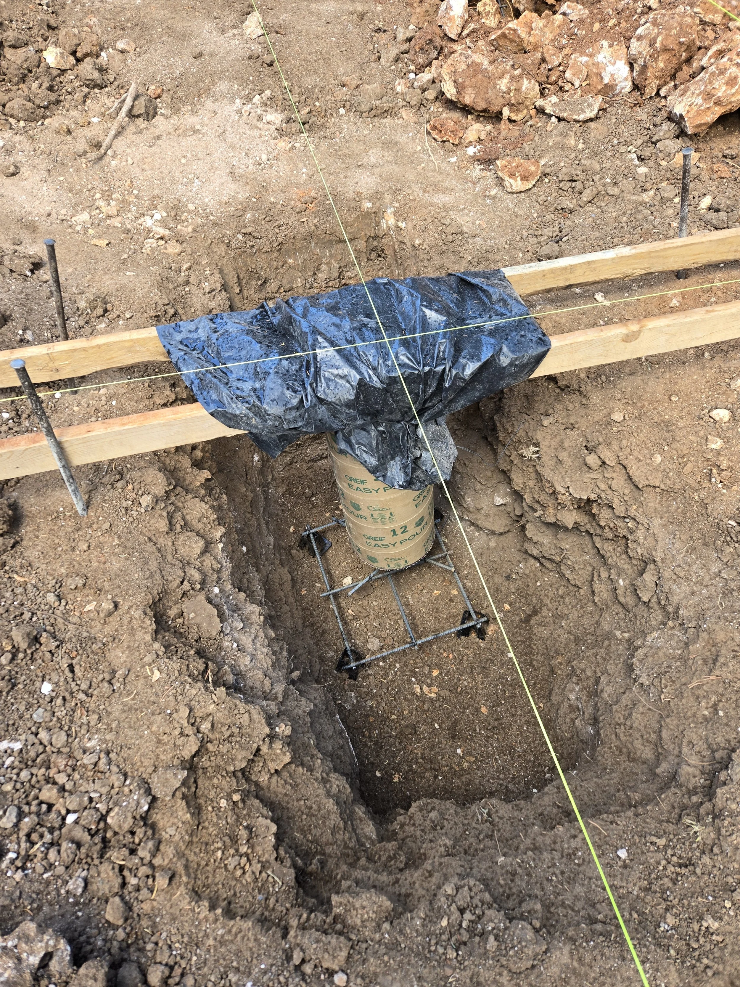 A construction site showing a hole in the ground with a plastic-covered pipe reinforced with rebar, wooden supports, and yellow string lines for leveling.