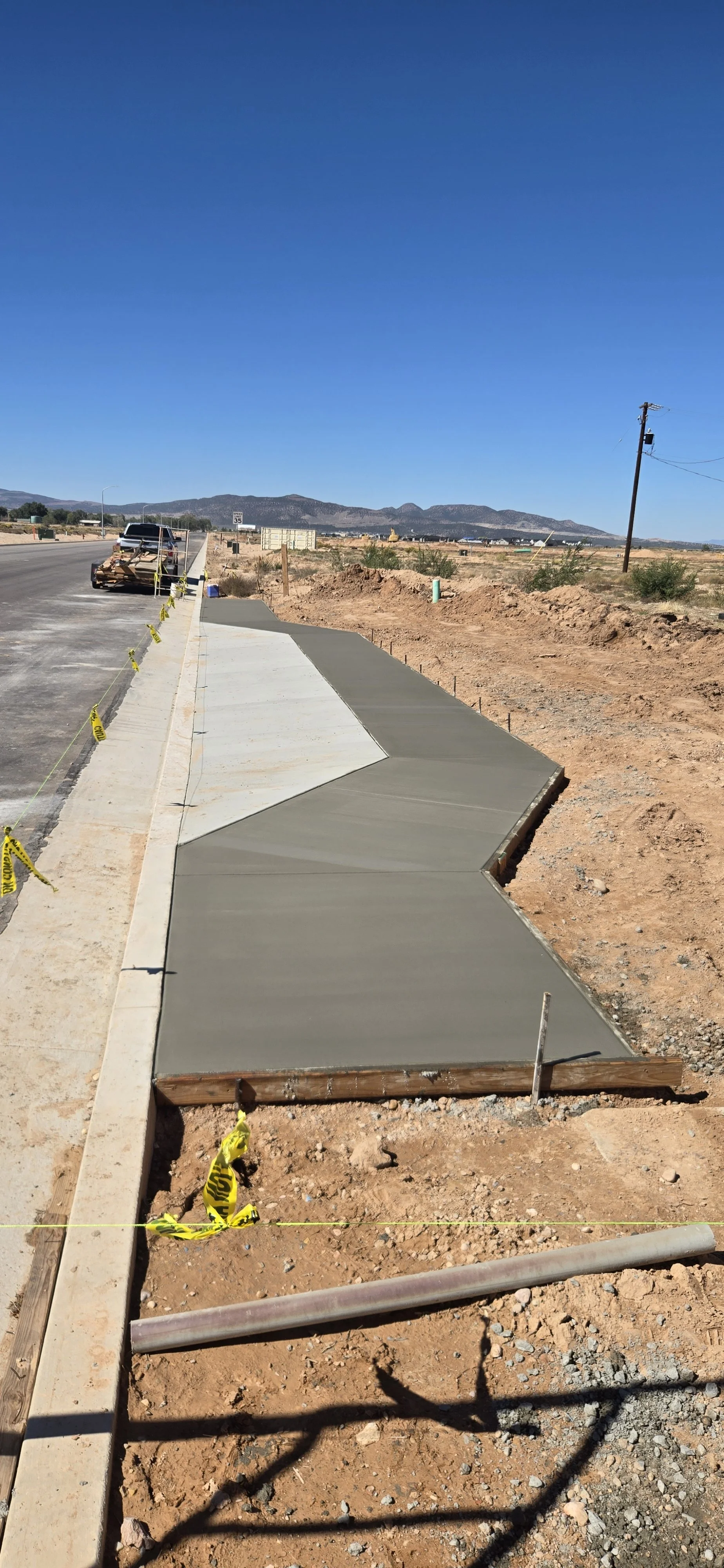 Newly poured concrete sidewalk under construction along a dusty, open area with mountains in the background, a utility pole, and cloudy sky.