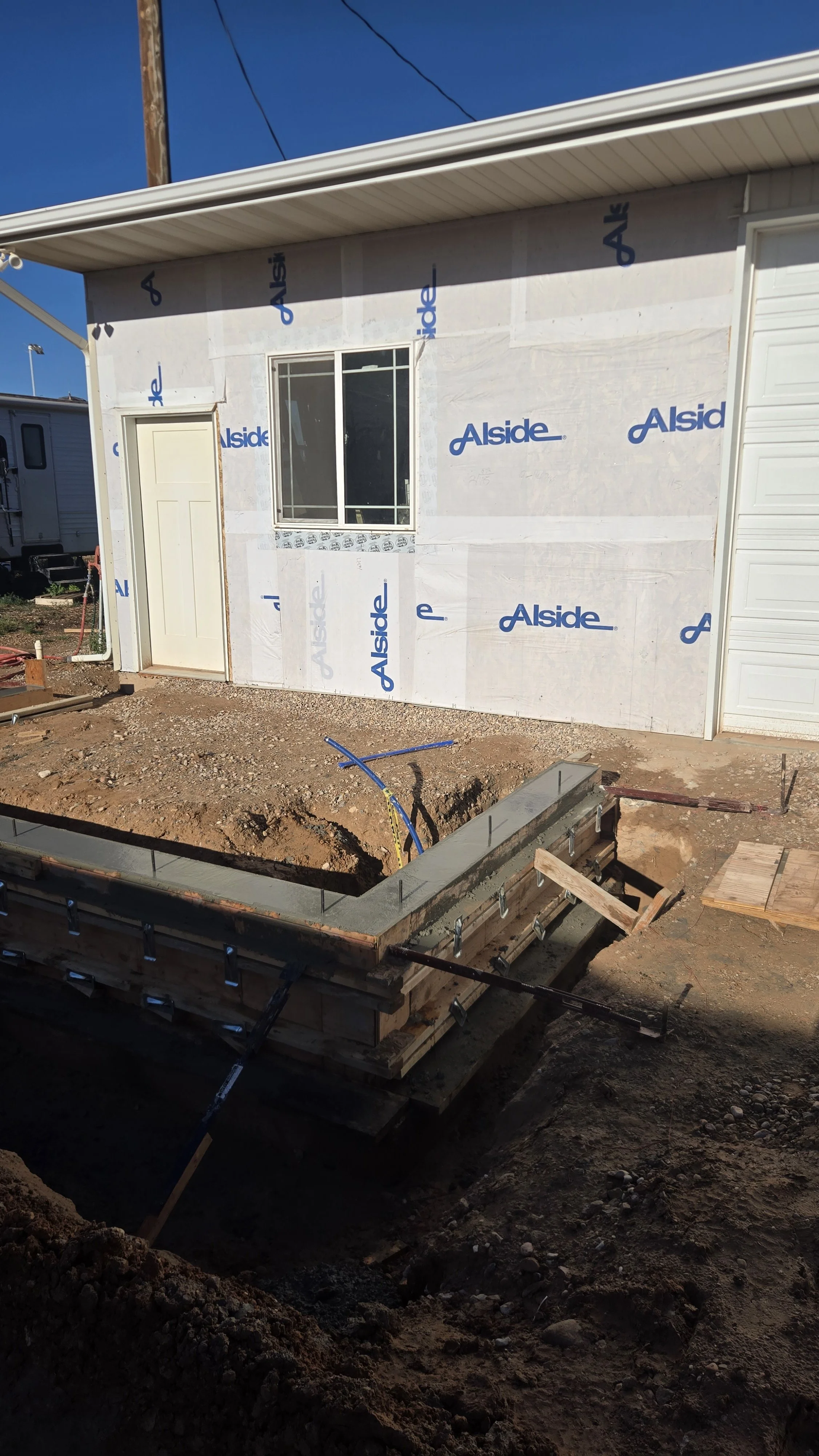 Construction site with house in early framing stage, exterior walls covered with white sheathing marked 'Alside,' a window, a door, and an open foundation with rebar and wooden formwork for concrete.
